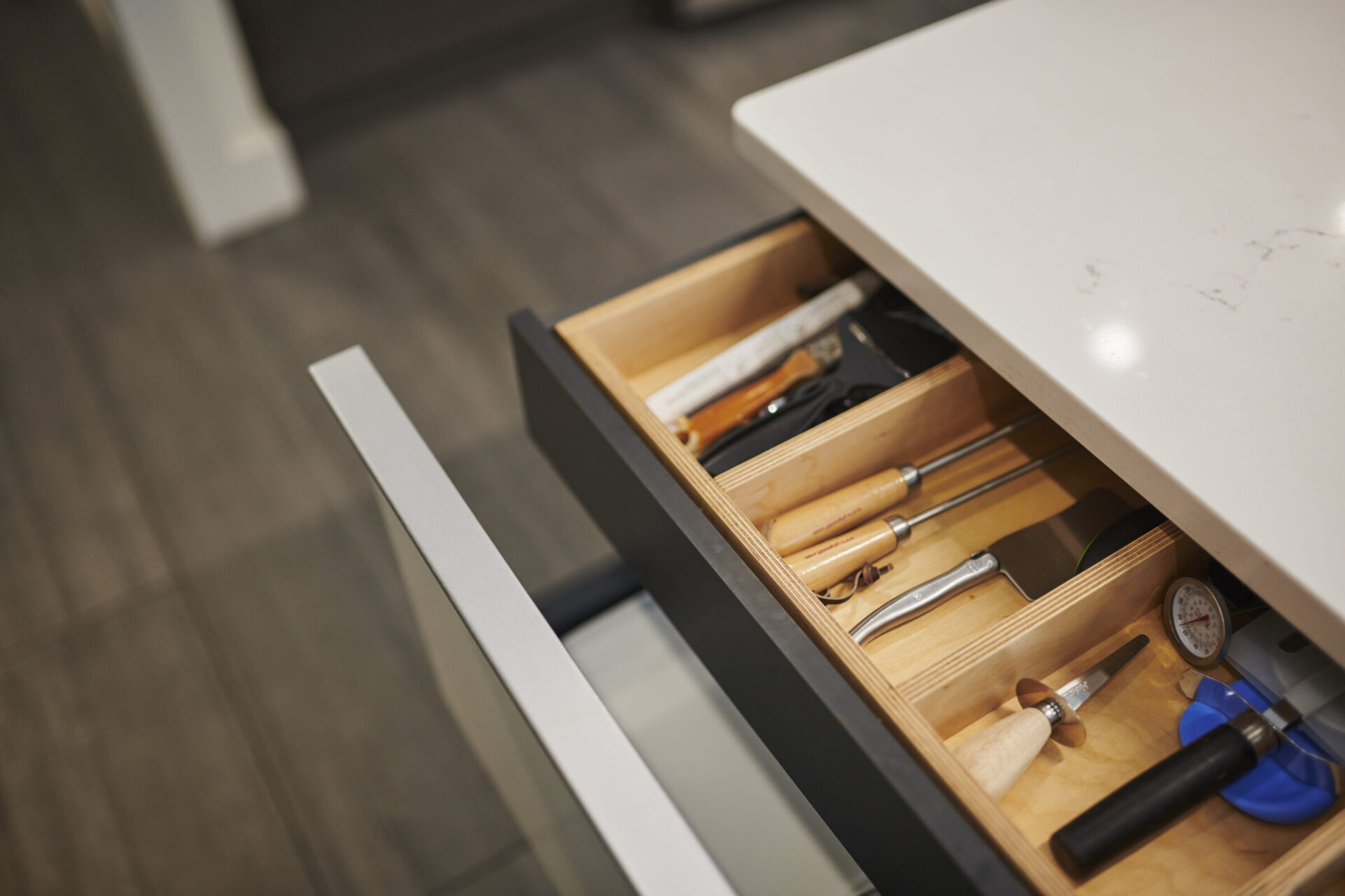 An open kitchen drawer reveals a collection of tools and utensils, including a hammer, chisels, and a tape measure, with a white countertop above.