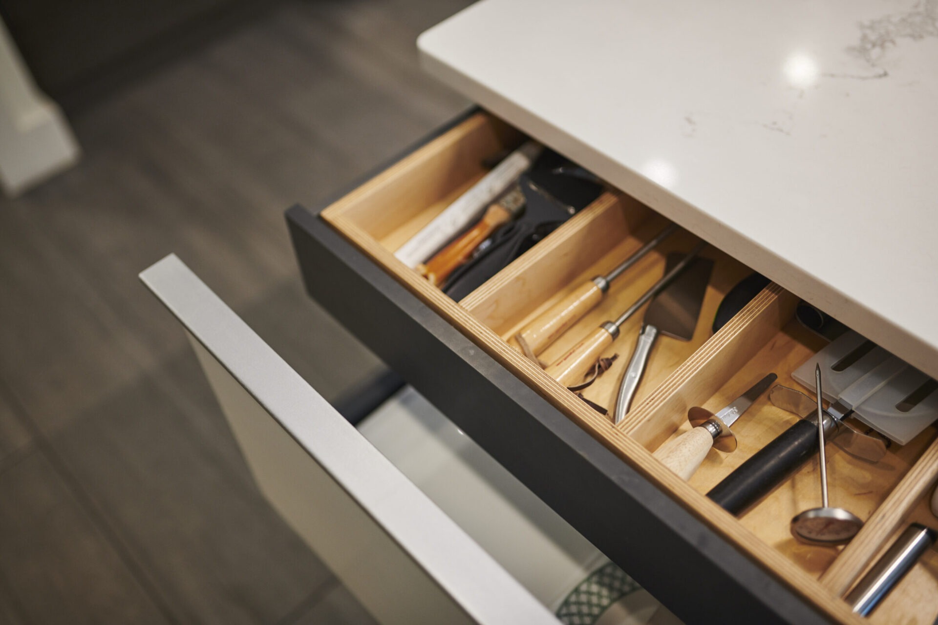 An open kitchen drawer reveals an organized array of utensils, including knives and a cheese slicer, neatly arranged in wooden dividers on a countertop.