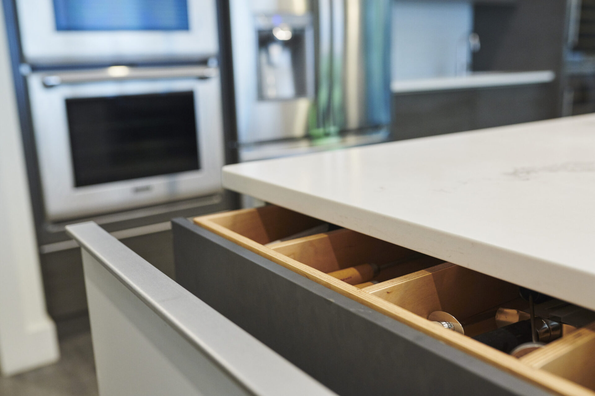 Modern kitchen interior showing a close-up of an open drawer with utensils, a countertop, stainless steel oven, and refrigerator in soft focus background.