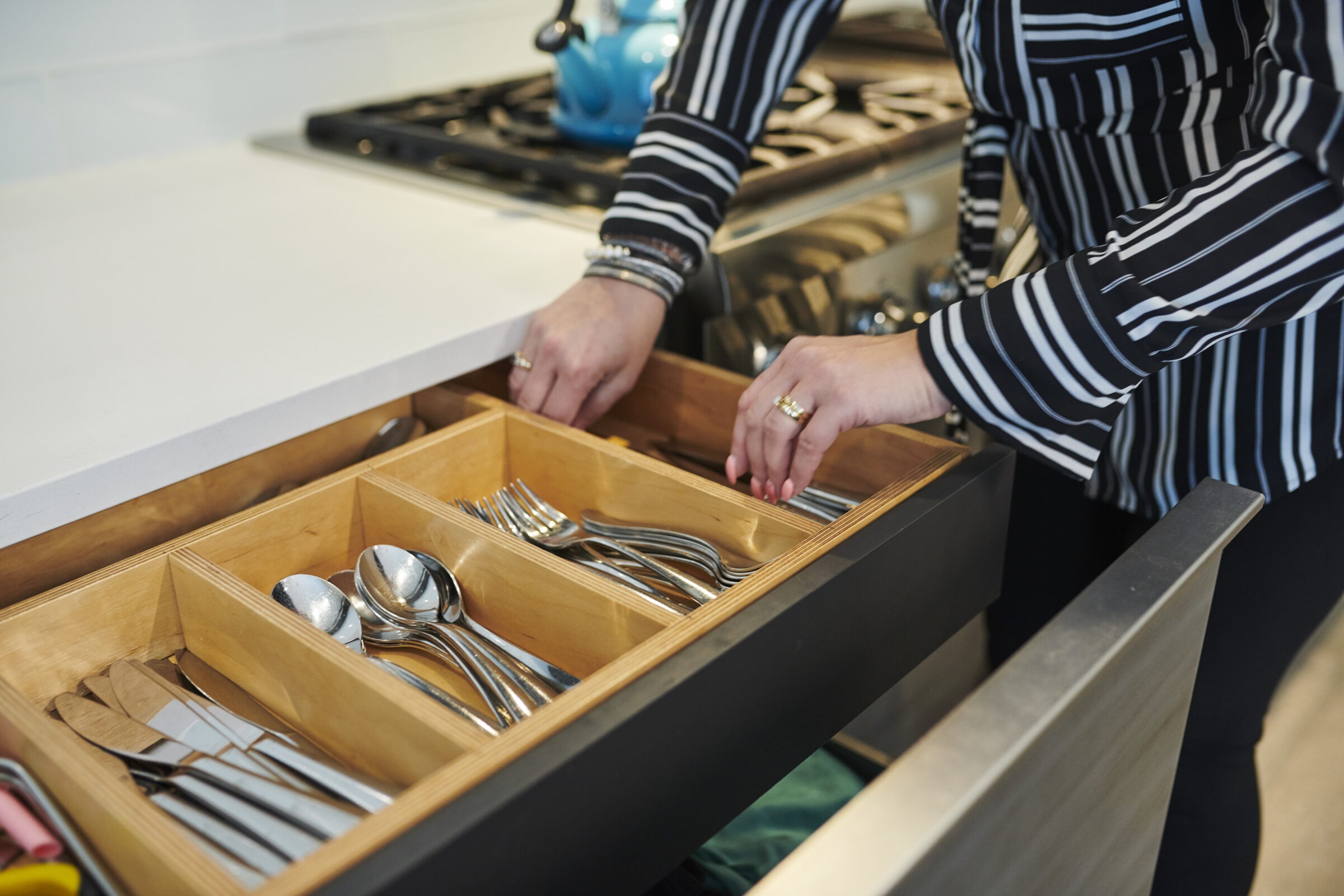 A person is organizing a kitchen drawer filled with cutlery including spoons and forks. The kitchen has modern appliances and a clean countertop.