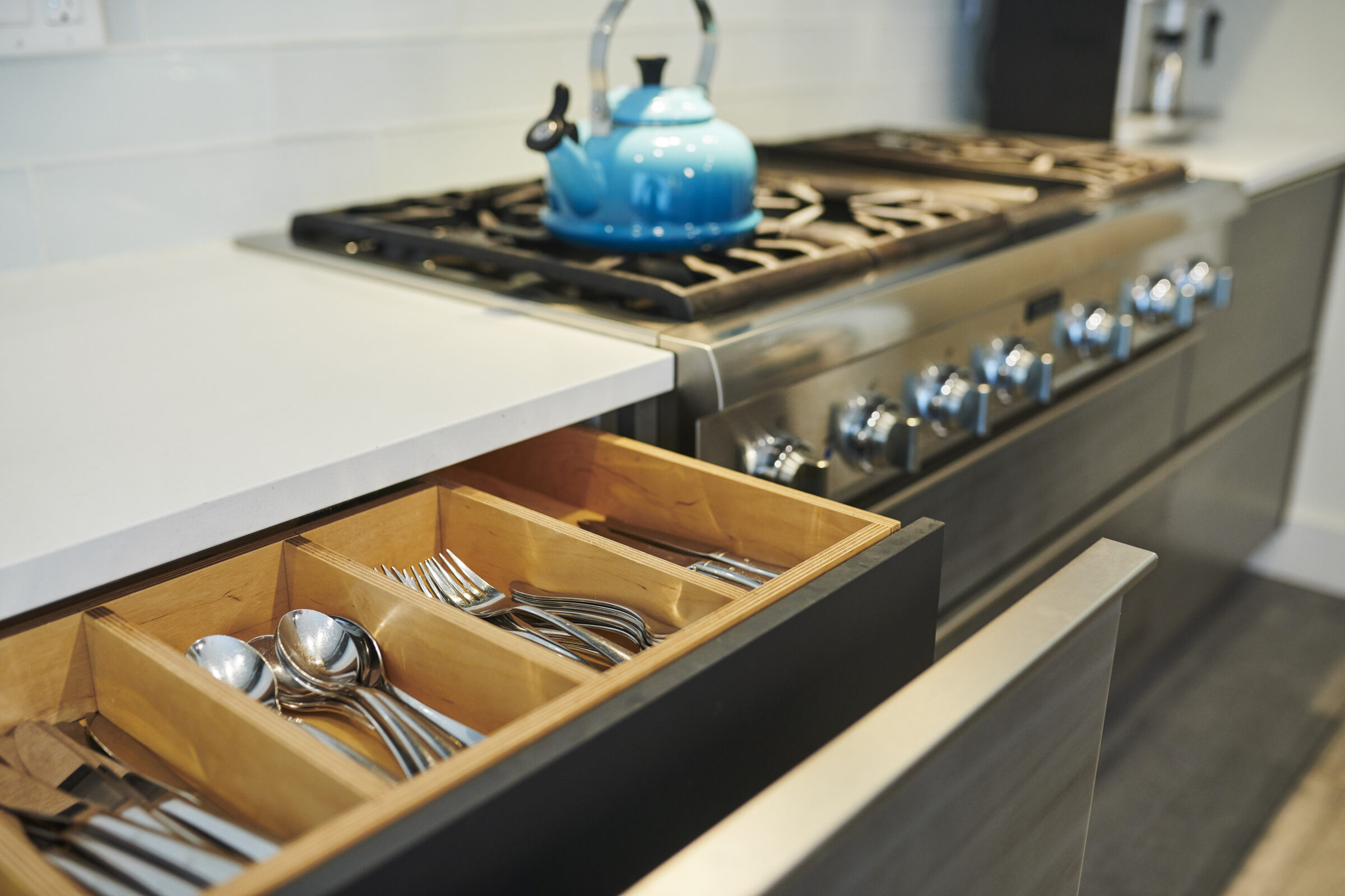 A modern kitchen with a blue kettle on a stainless steel gas stove, and an open drawer displaying organized silverware. Clean, contemporary design elements are evident.
