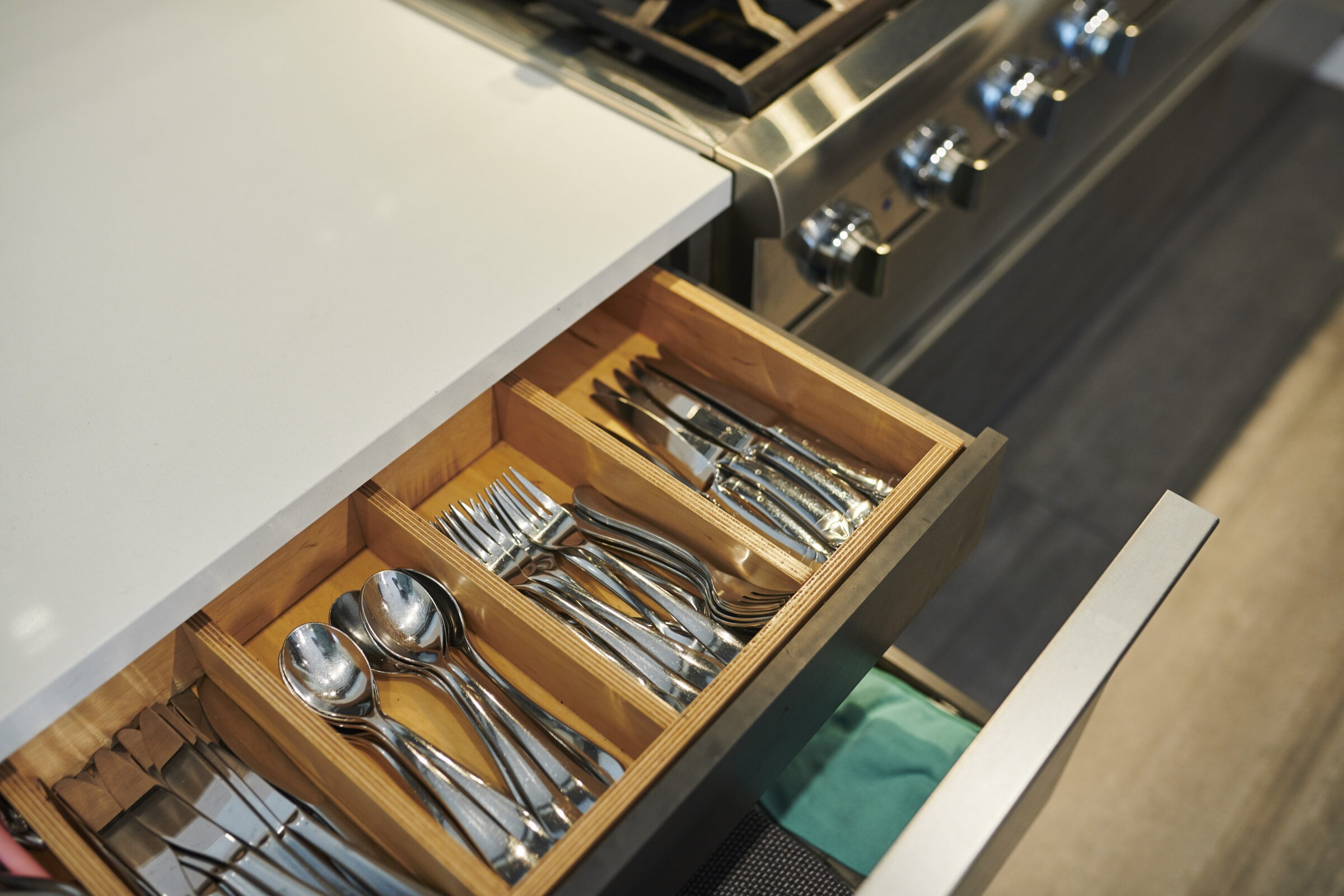 An open kitchen drawer reveals organized compartments holding silverware, including forks, knives, and spoons, adjacent to a modern stove with dials.