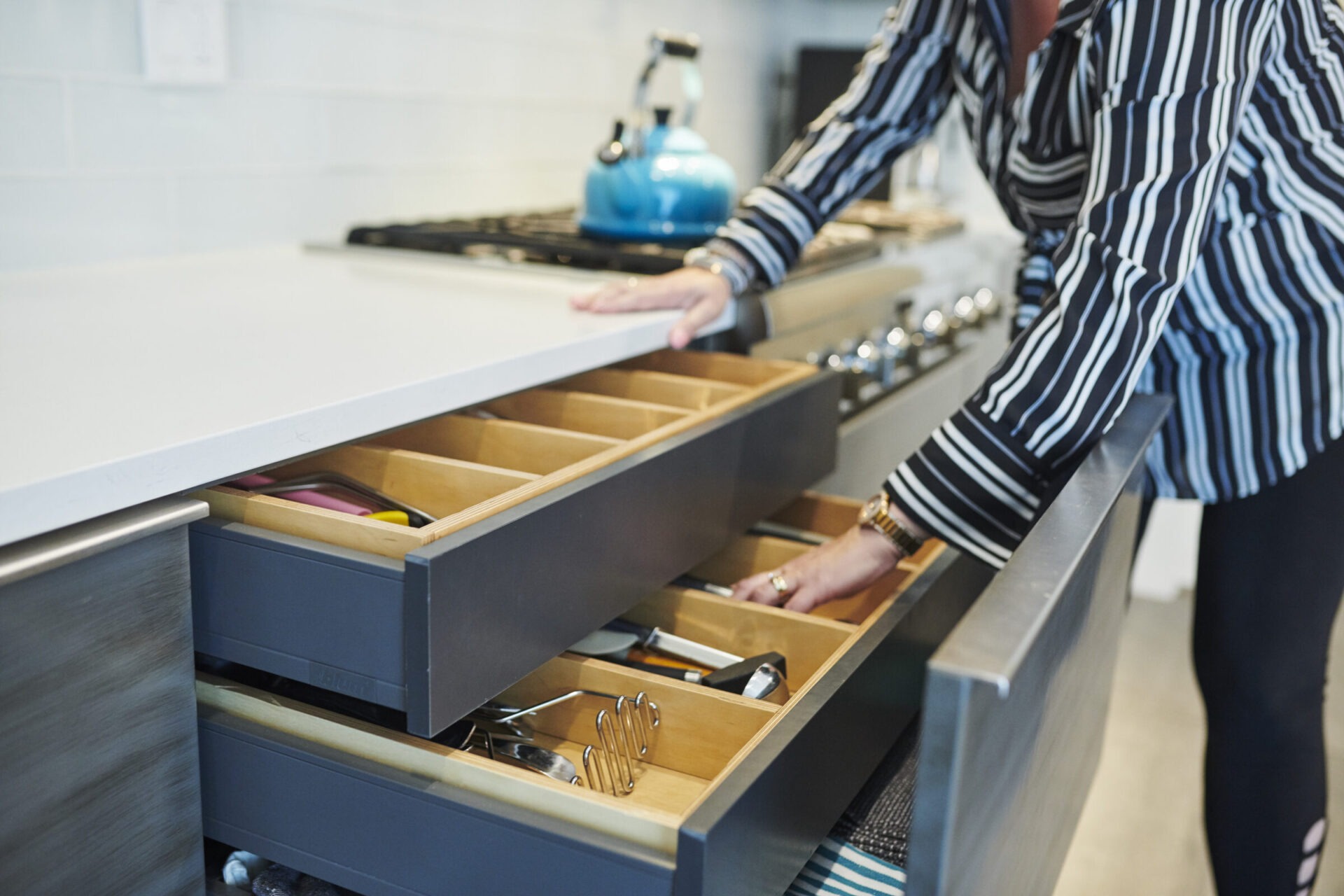 A person is standing at a modern kitchen counter, opening a drawer revealing organized utensils and cutlery. A blue kettle is on the countertop.