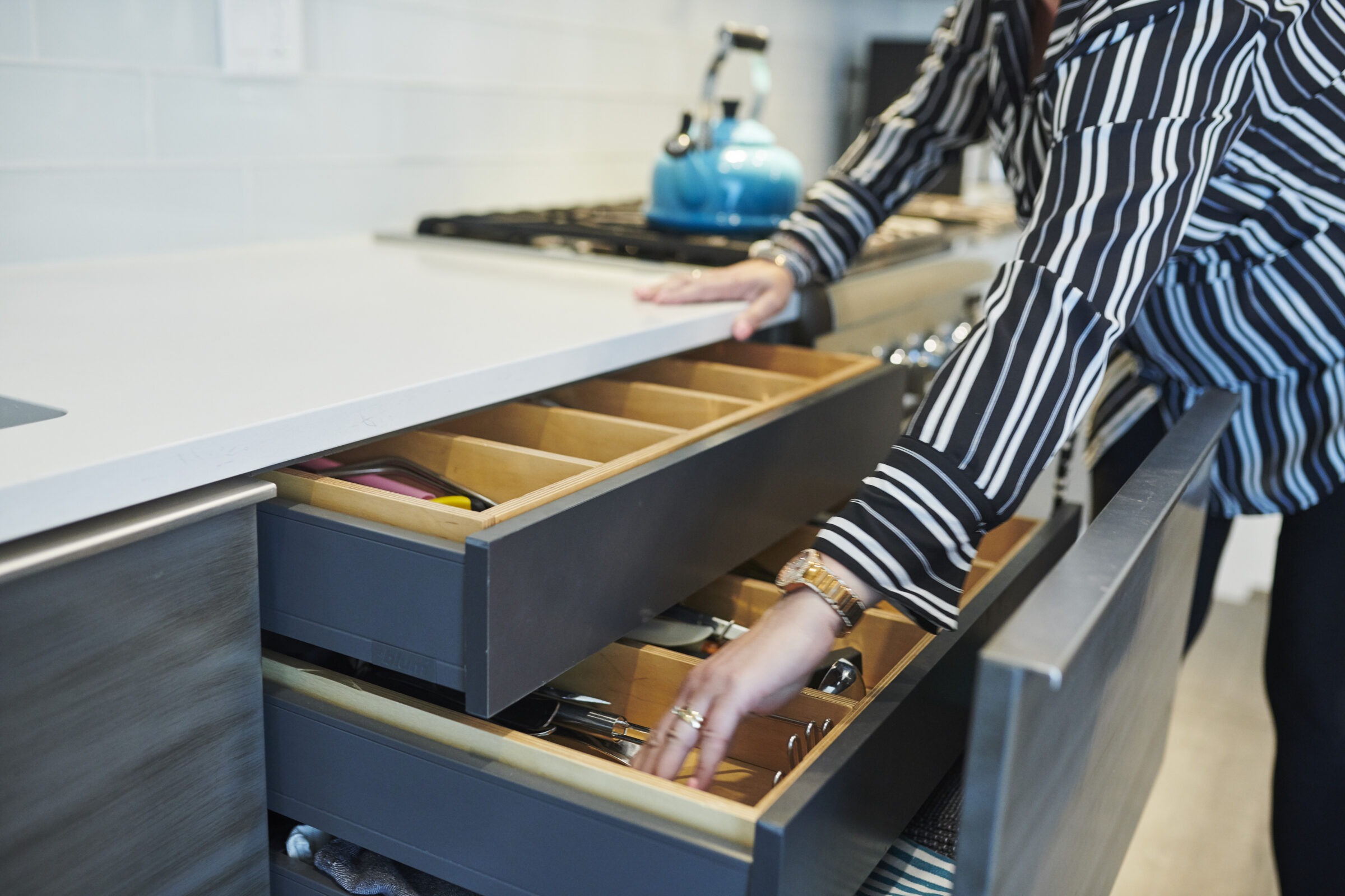 A person is standing by a modern kitchen counter, opening a drawer to reveal organized utensils. A teal kettle rests on the countertop.
