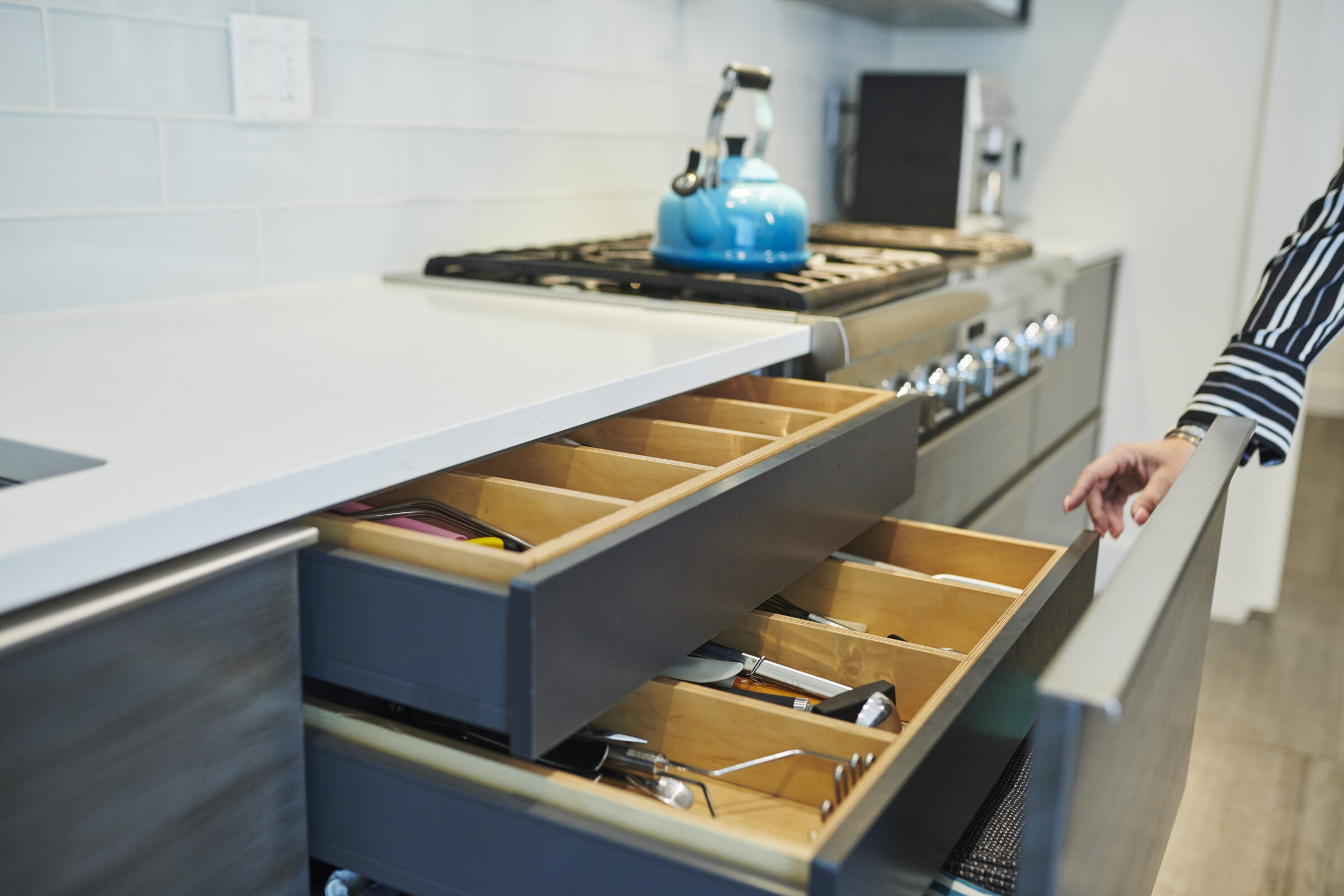 A modern kitchen with a person opening organized drawers containing utensils. There's a blue kettle on the stove and white countertops.