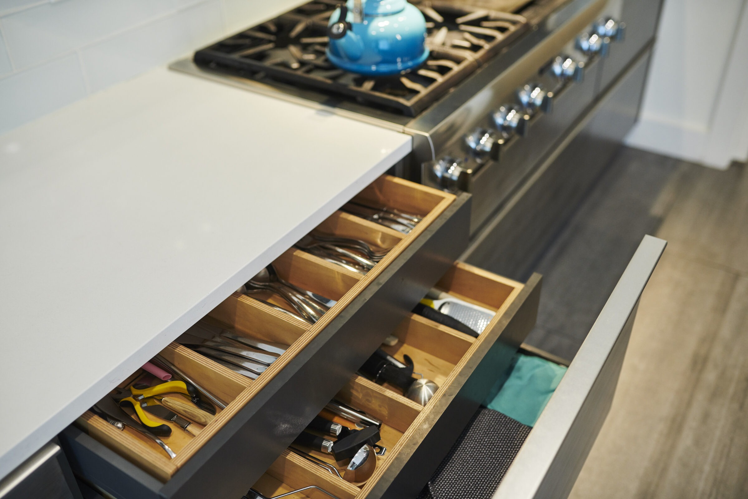 A modern kitchen with open drawers showing organized utensils. A gas stove with stainless steel finish and blue kettle on top.