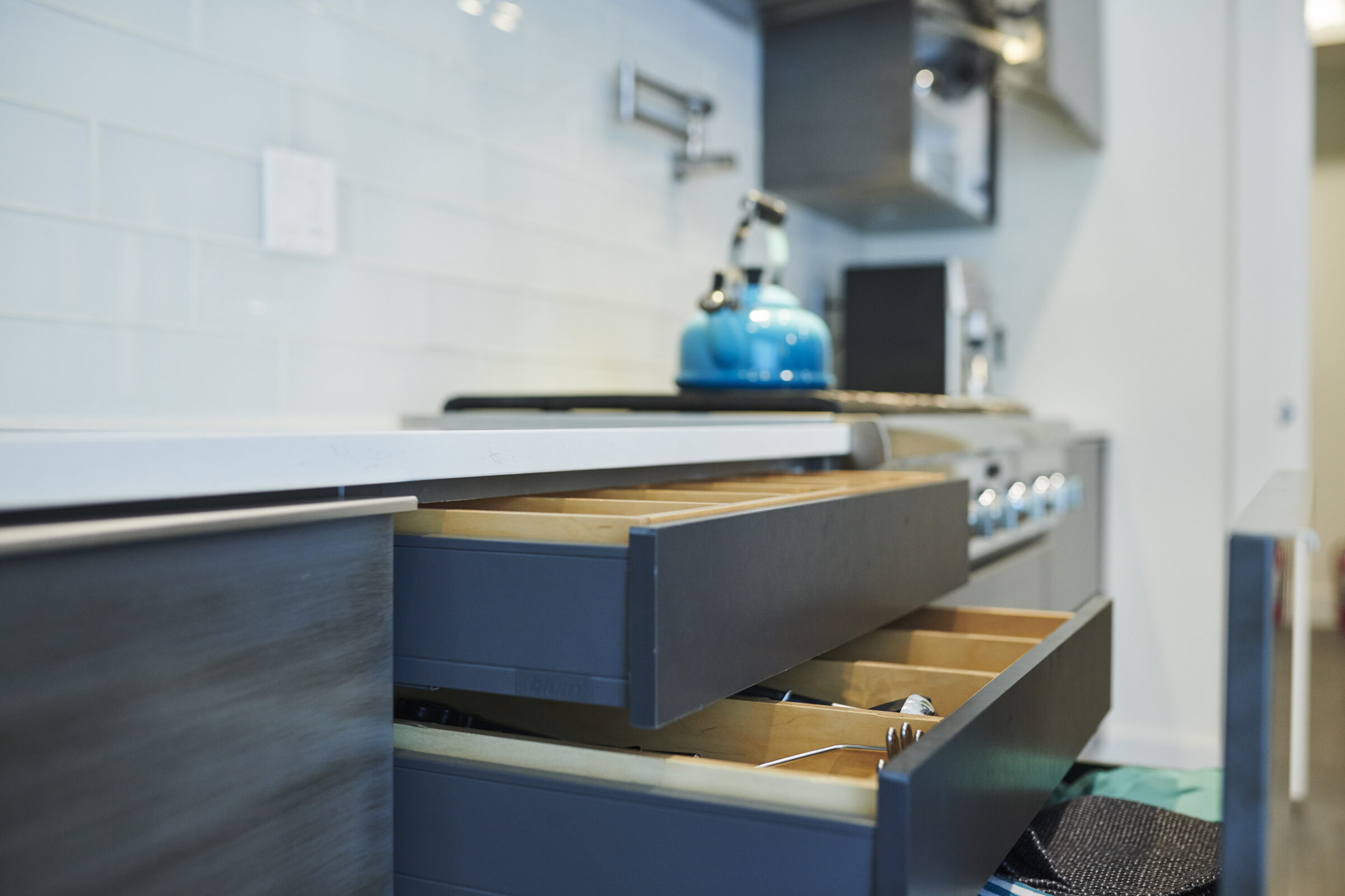 A modern kitchen with open drawers revealing utensils, a blue kettle on the countertop, and a blurred background featuring a stove and cabinets.