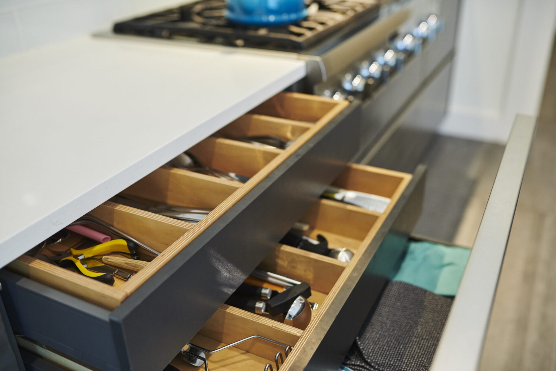 A modern kitchen with a white countertop, open wooden drawers displaying utensils and tools, and a blue pot on a gas stove.