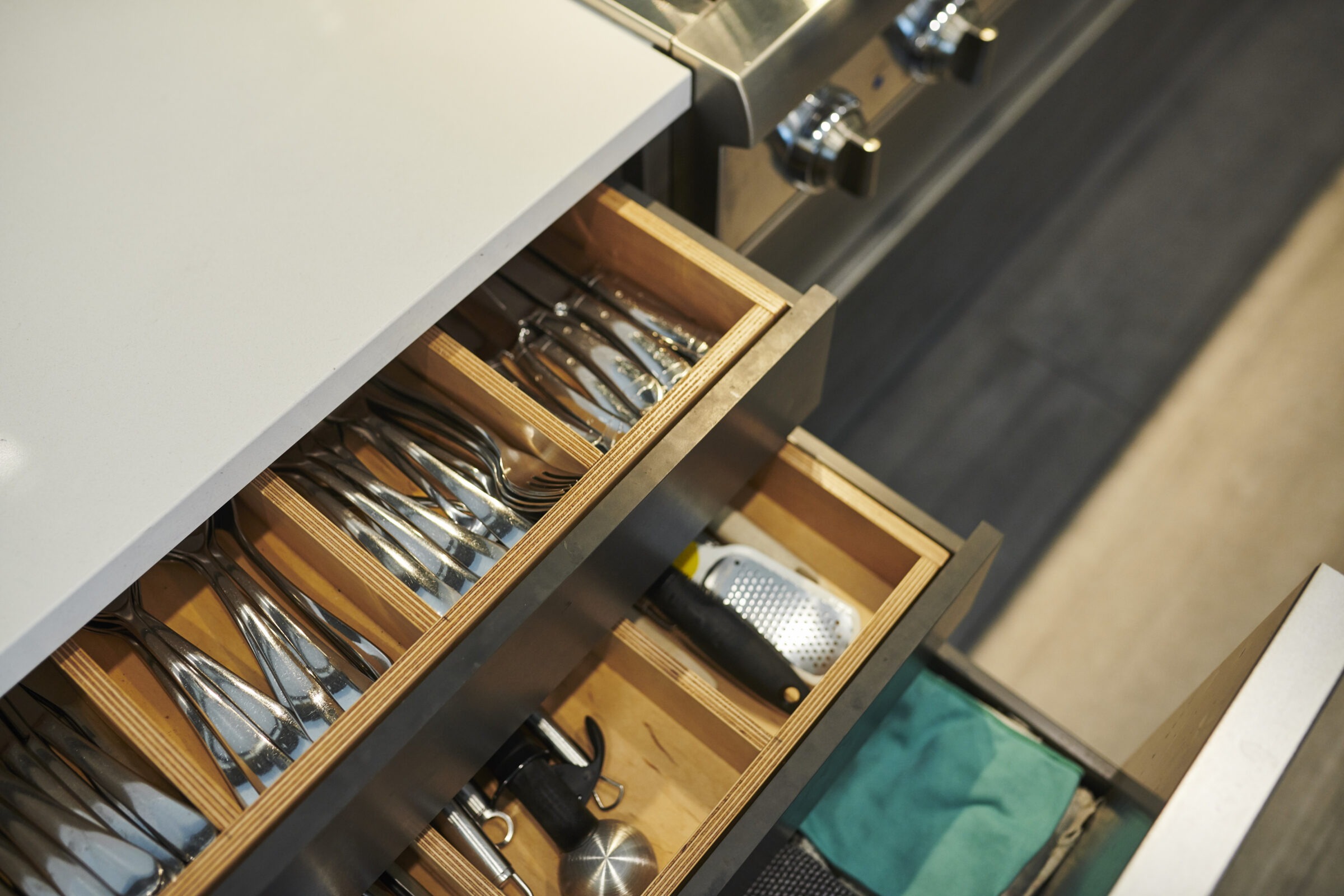 An open kitchen drawer reveals neatly organized cutlery and utensils. The modern design features wood separators and a mix of stainless steel tools.