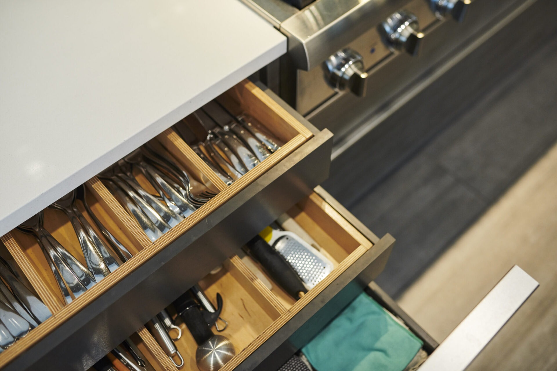 A modern kitchen drawer partially open, revealing organized utensils like forks and knives, next to a stainless steel oven with knobs.