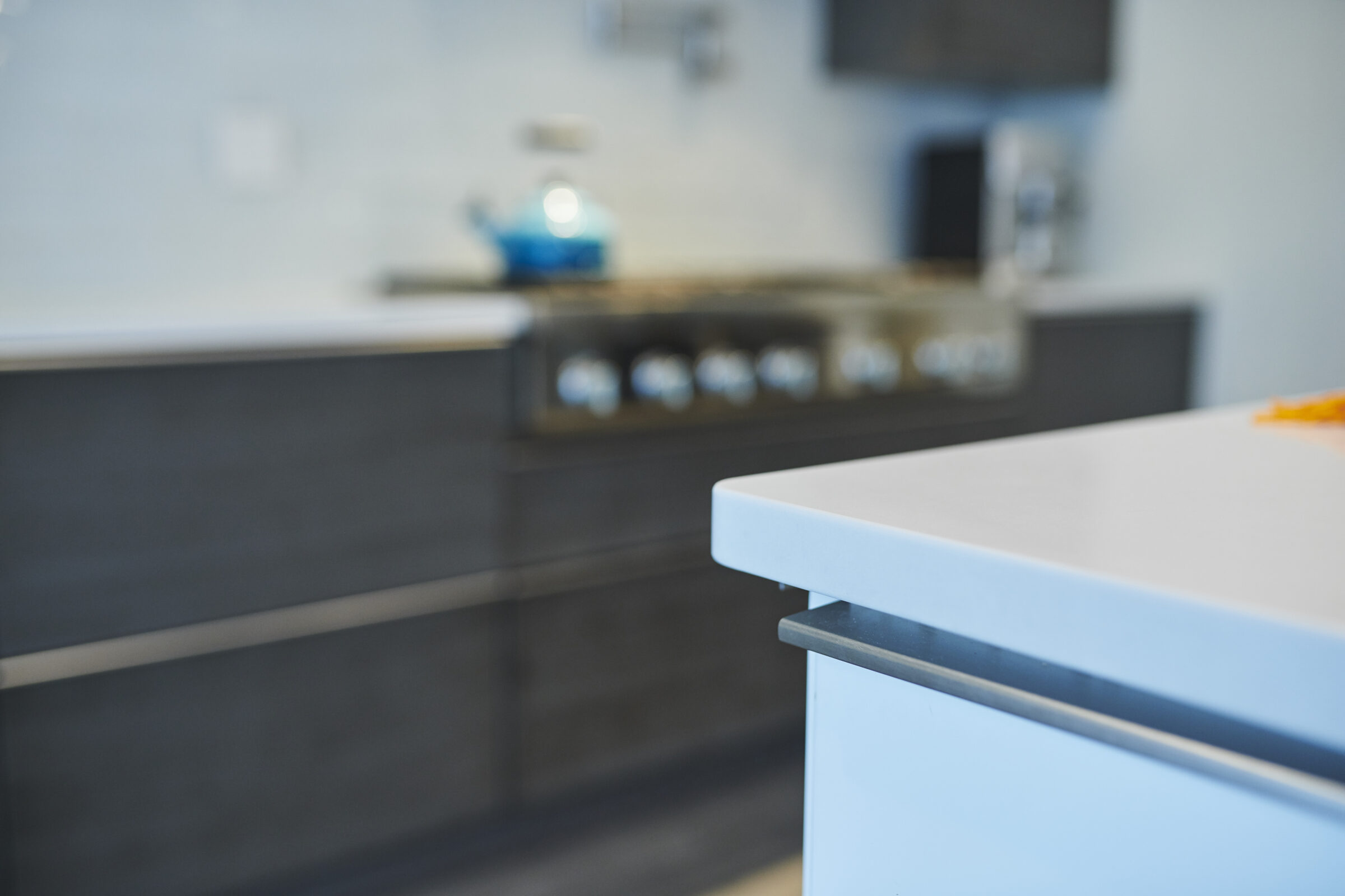 Modern kitchen interior focused on a white countertop edge with blurred background of cabinets, stove, and blue decor accents. Clean, contemporary design.