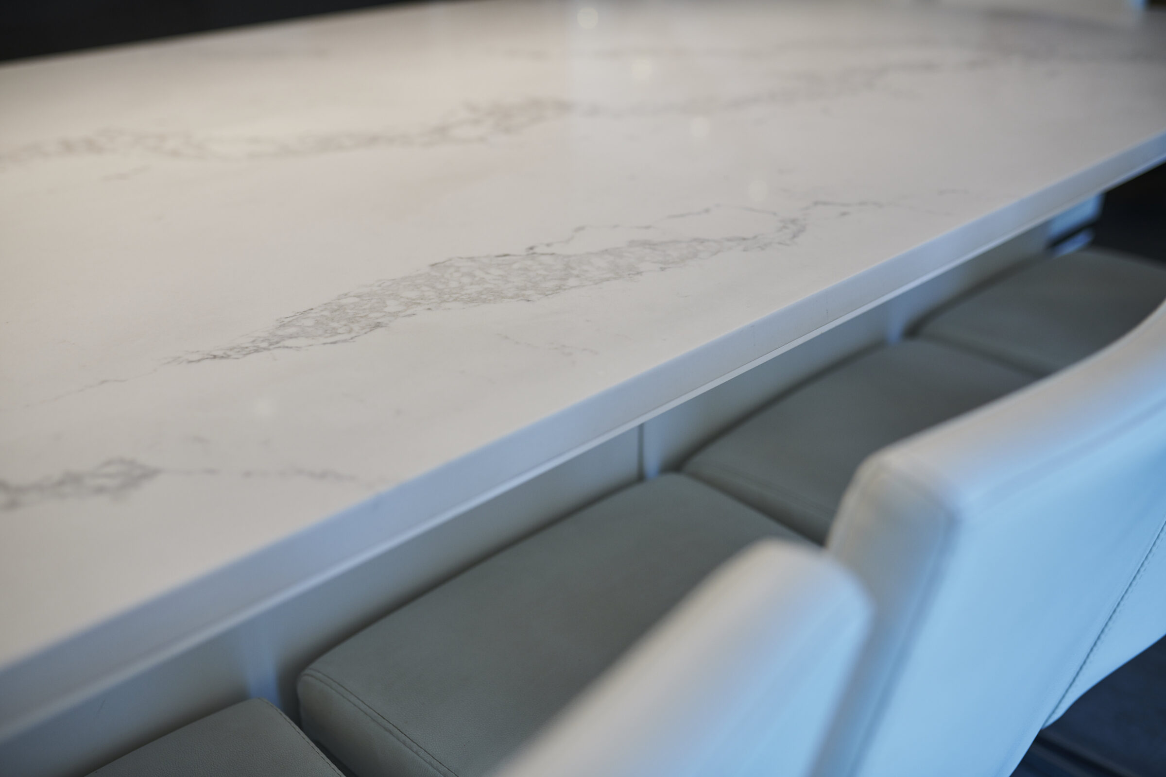 The image shows a close-up of a white marble countertop above a contemporary style seating area with padded grey chairs. No people are visible.