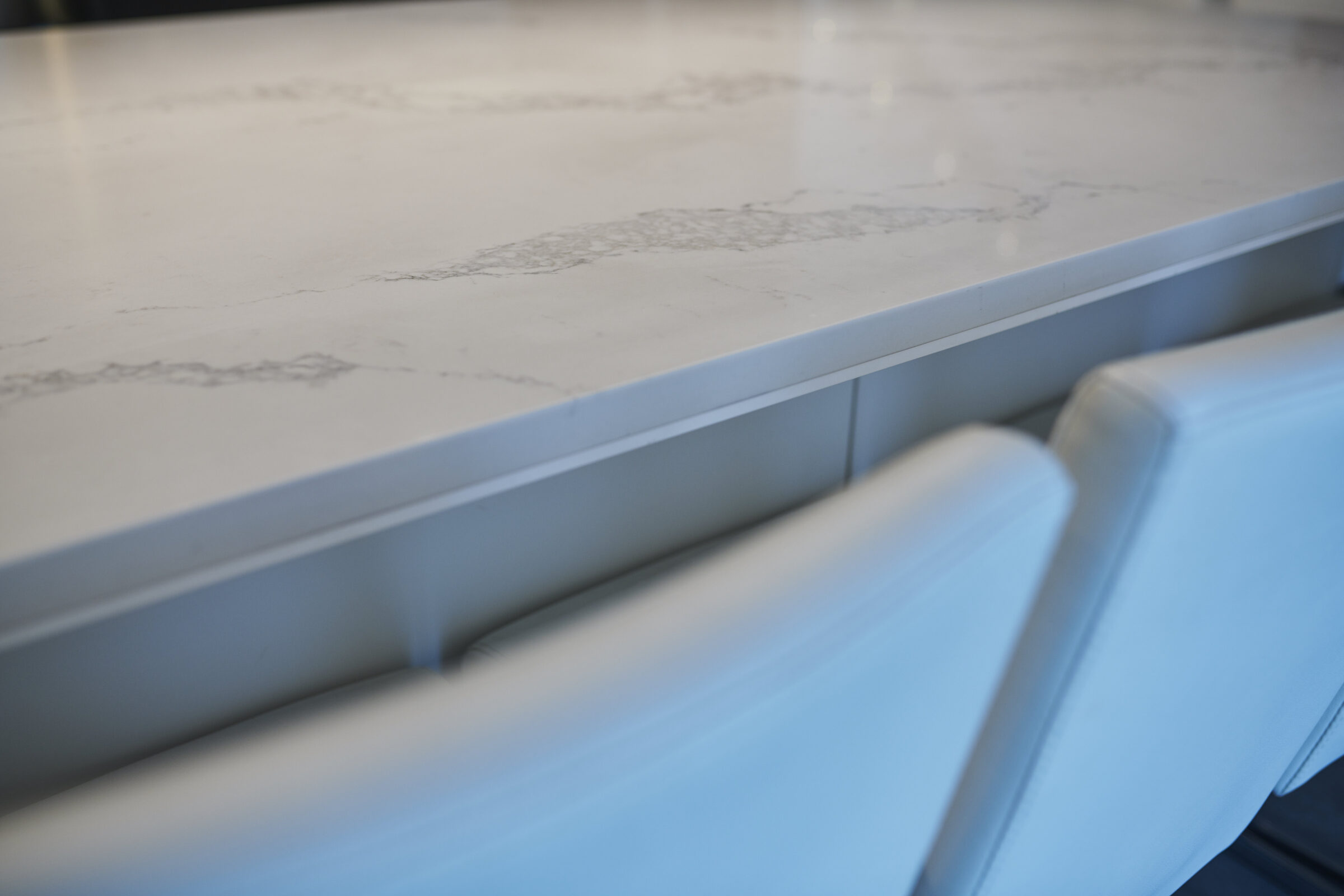 The image shows a close-up of a marble countertop with a vein pattern above a white drawer, next to a blurred blue-upholstered chair or sofa.