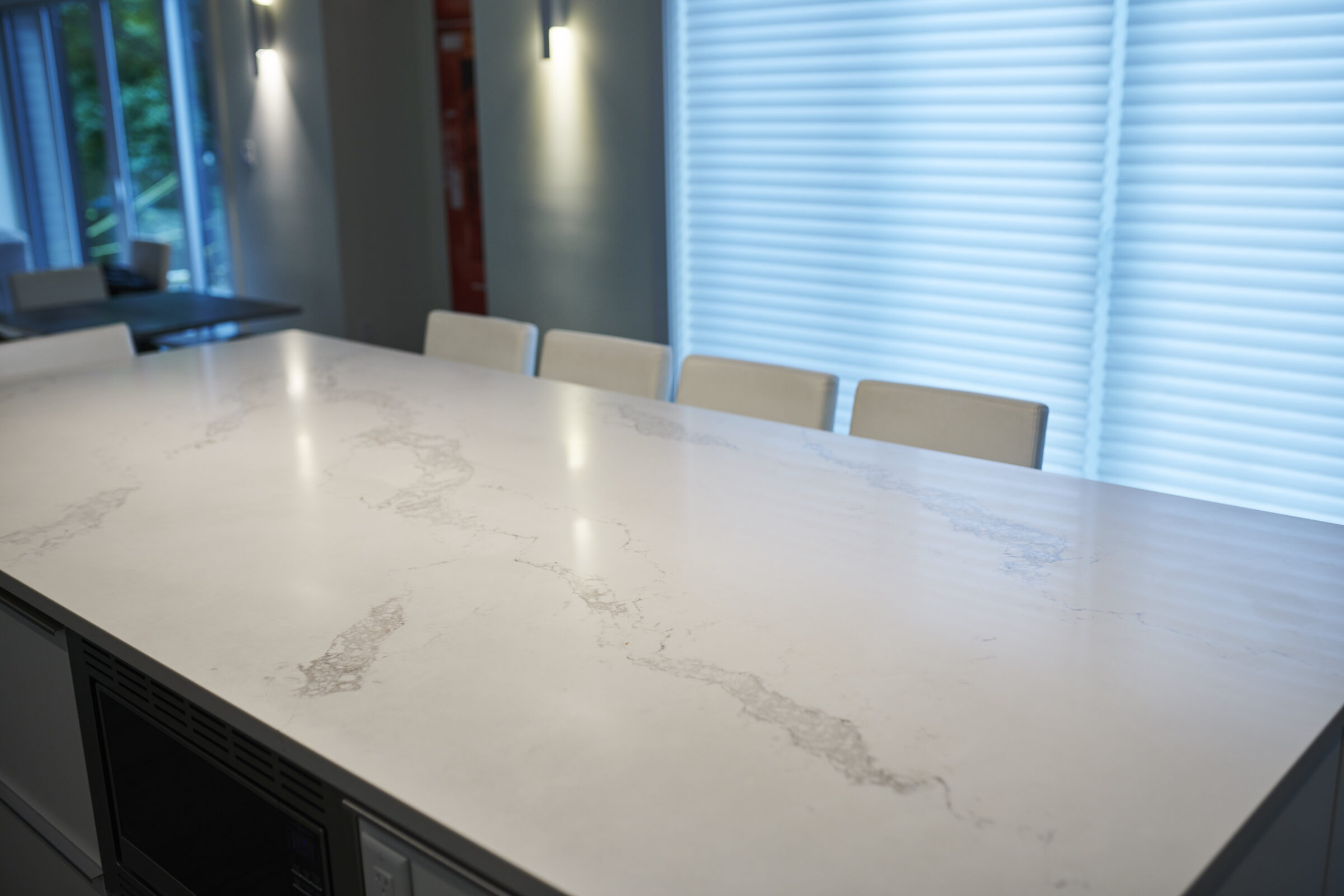 A modern kitchen interior with a large marble-patterned island countertop, white chairs, and venetian blinds covering windows in the background.