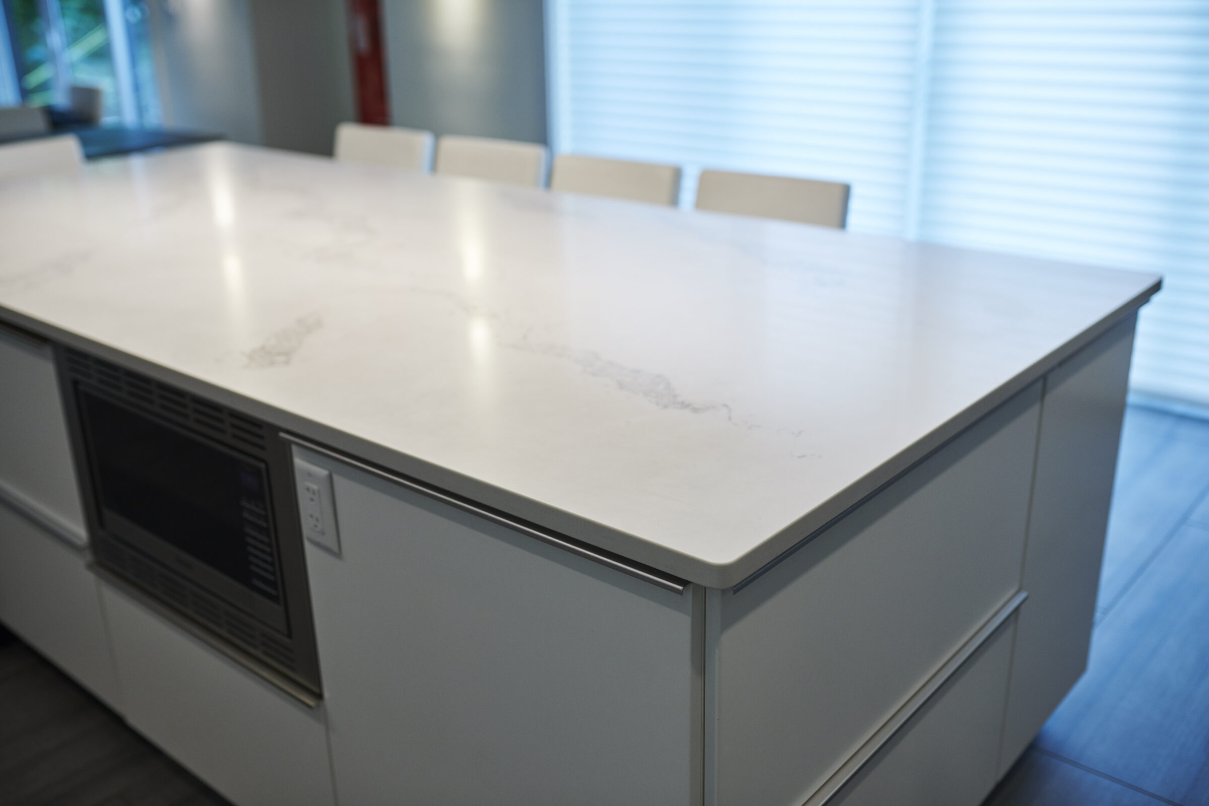 A modern kitchen island featuring a white marble countertop with a subtle vein pattern, white cabinets, and a built-in microwave. Sunshine filters through venetian blinds.