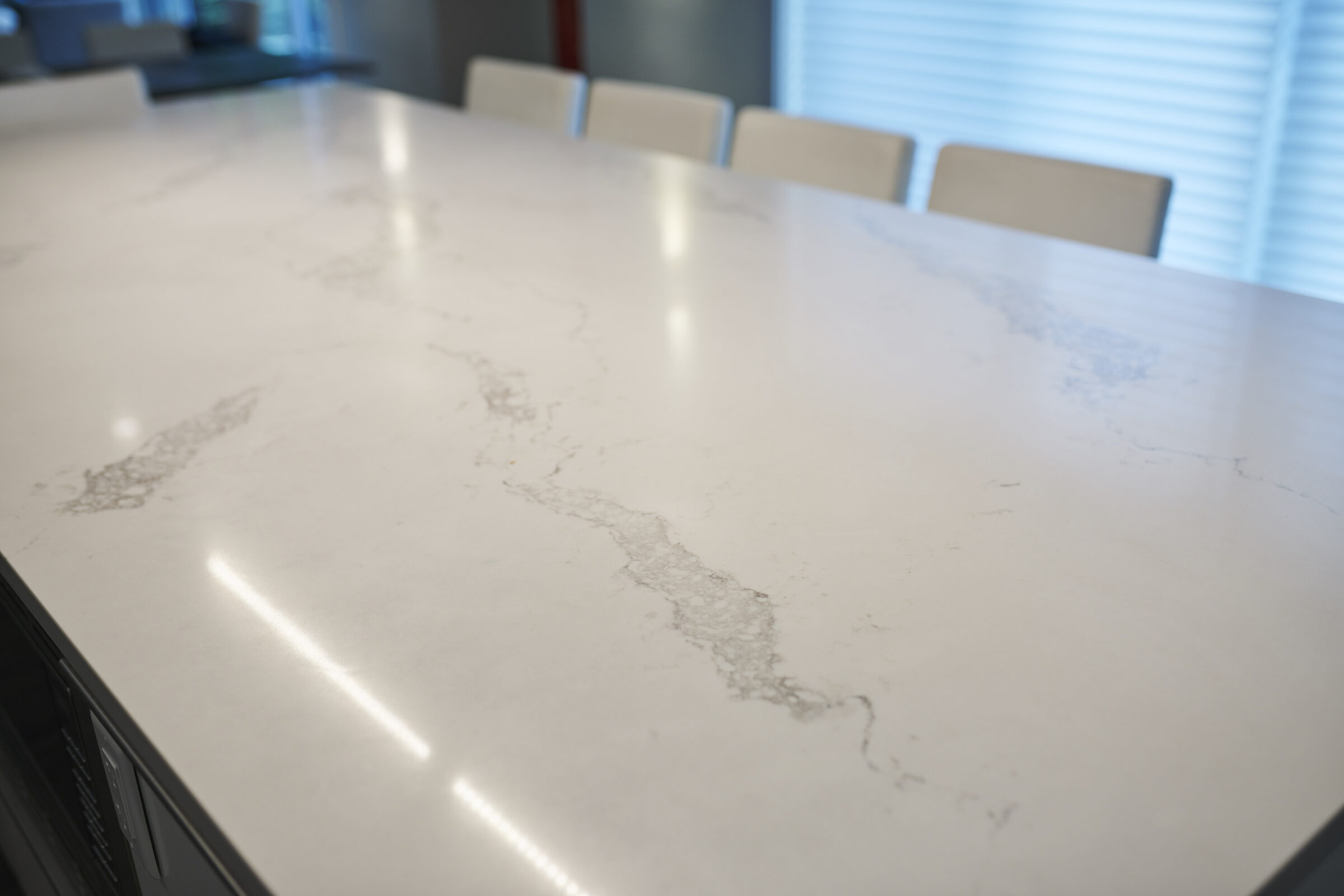 A modern kitchen with a reflective white marble countertop and six chairs, under natural light with a glimpse of blinds in the background.