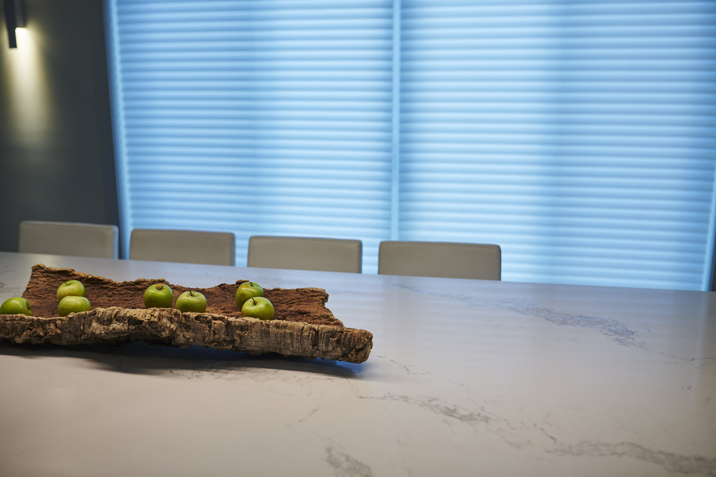 A modern kitchen scene with a marble countertop, unique wooden centerpiece holding green apples, chairs, and blue blinds softly illuminating the space.