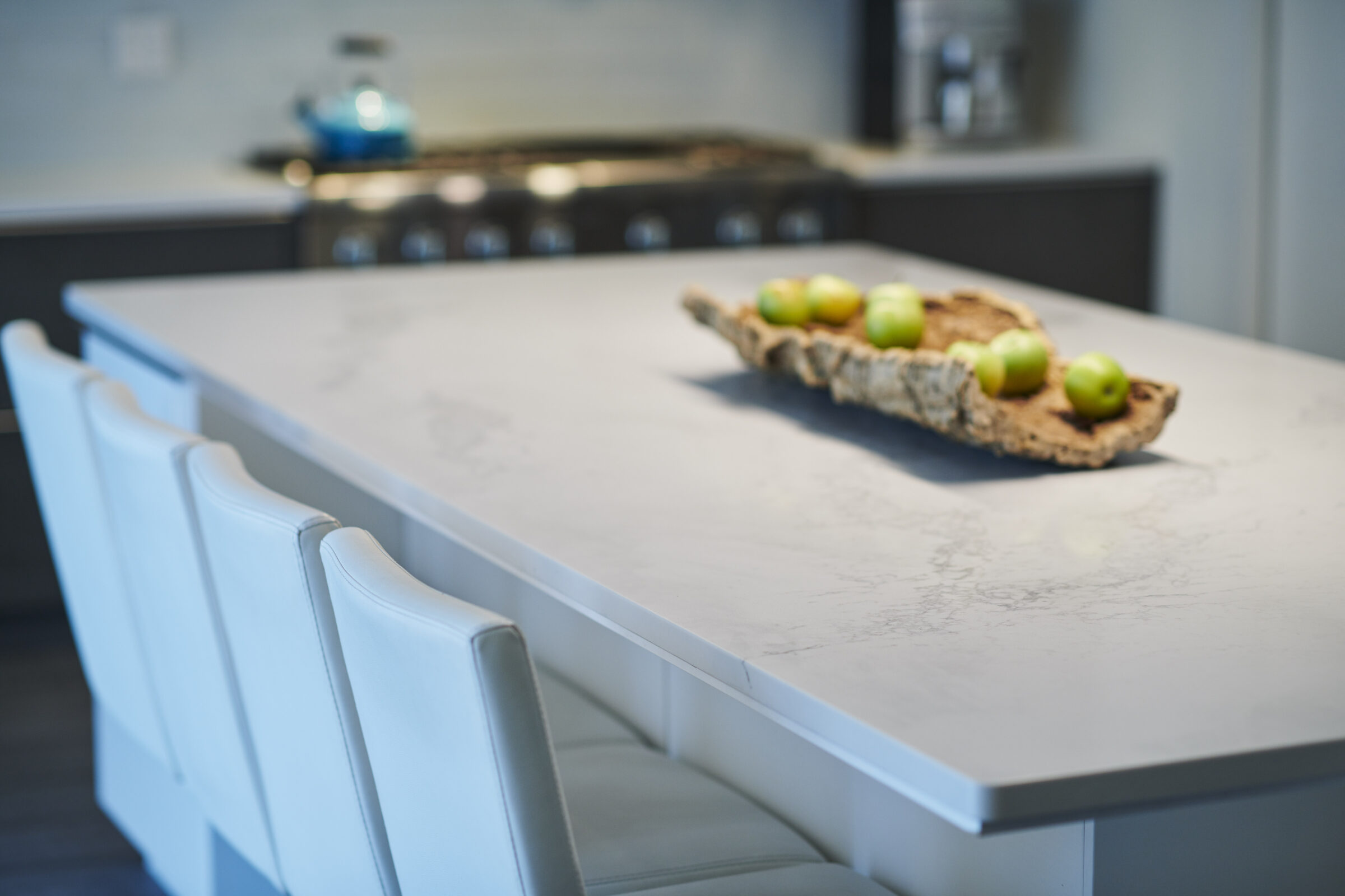 Modern kitchen with a white marble countertop, stylish white chairs, and a decorative wooden tray holding green apples, emphasizing clean, minimalist design.