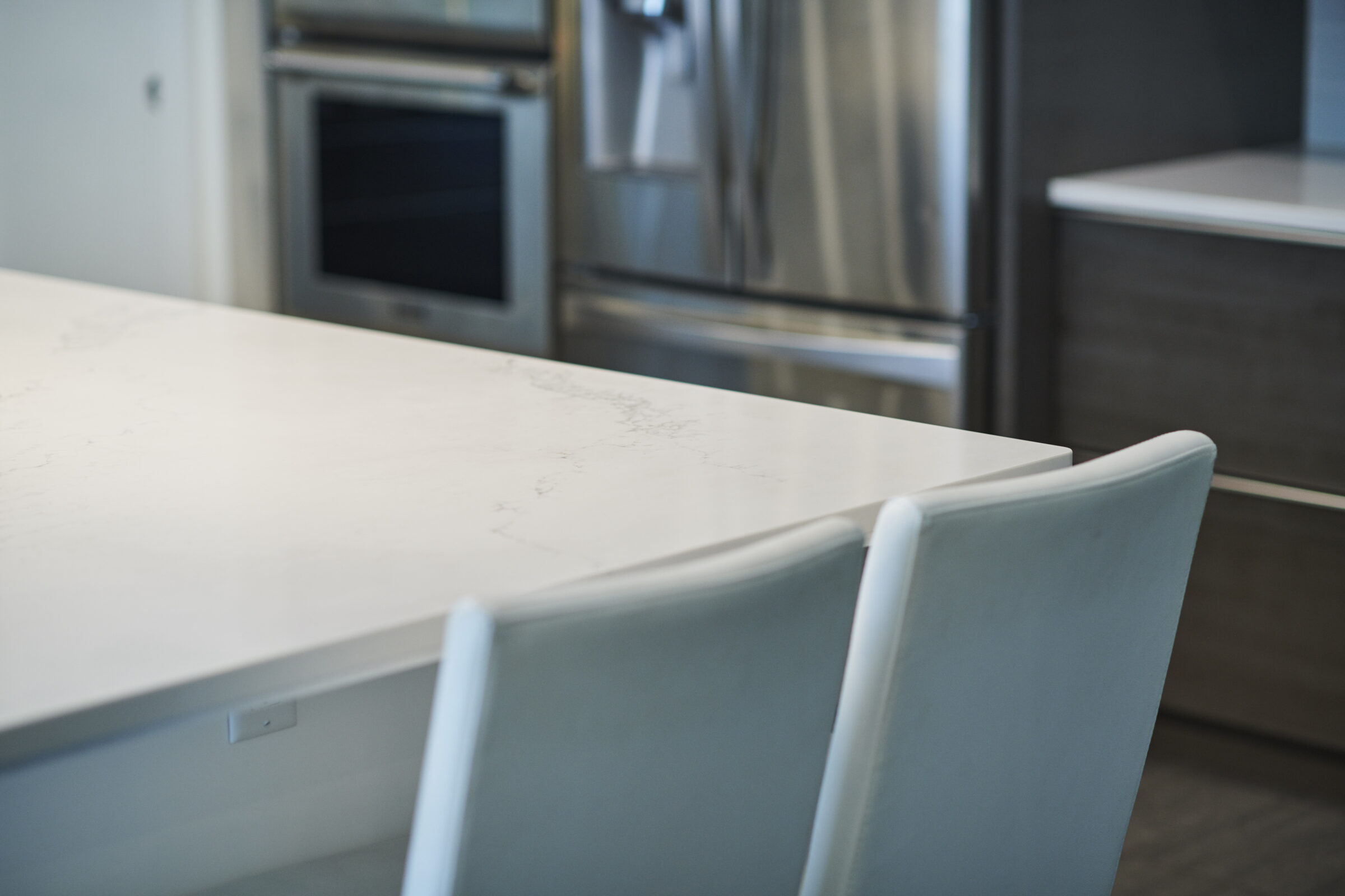 A modern kitchen with a white marble countertop, white chairs, stainless steel appliances, and contemporary cabinetry, with a focus on clean lines and minimalism.