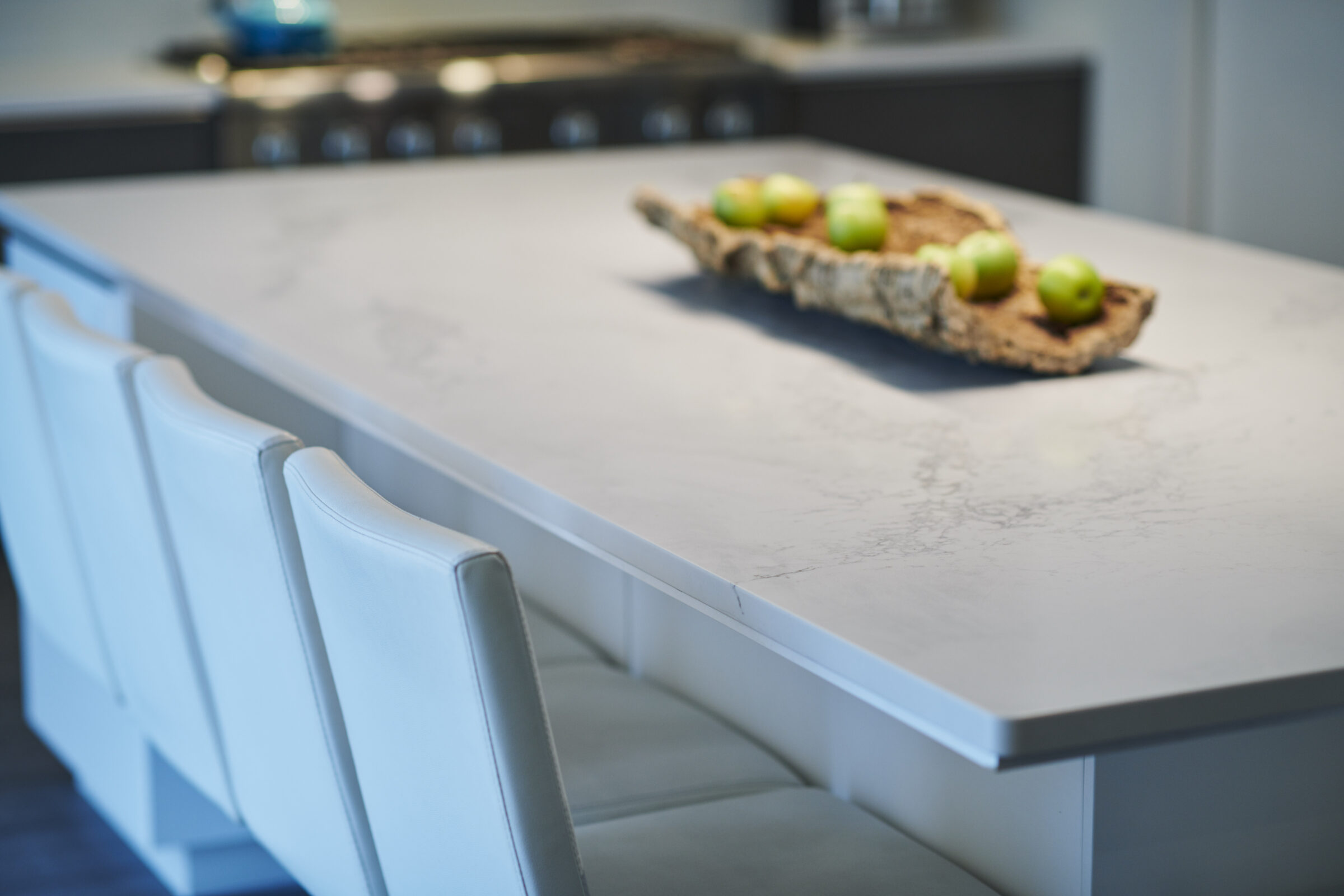 This image features a modern kitchen with a white marble countertop, elegant white chairs, and green apples arranged in a rustic tray.
