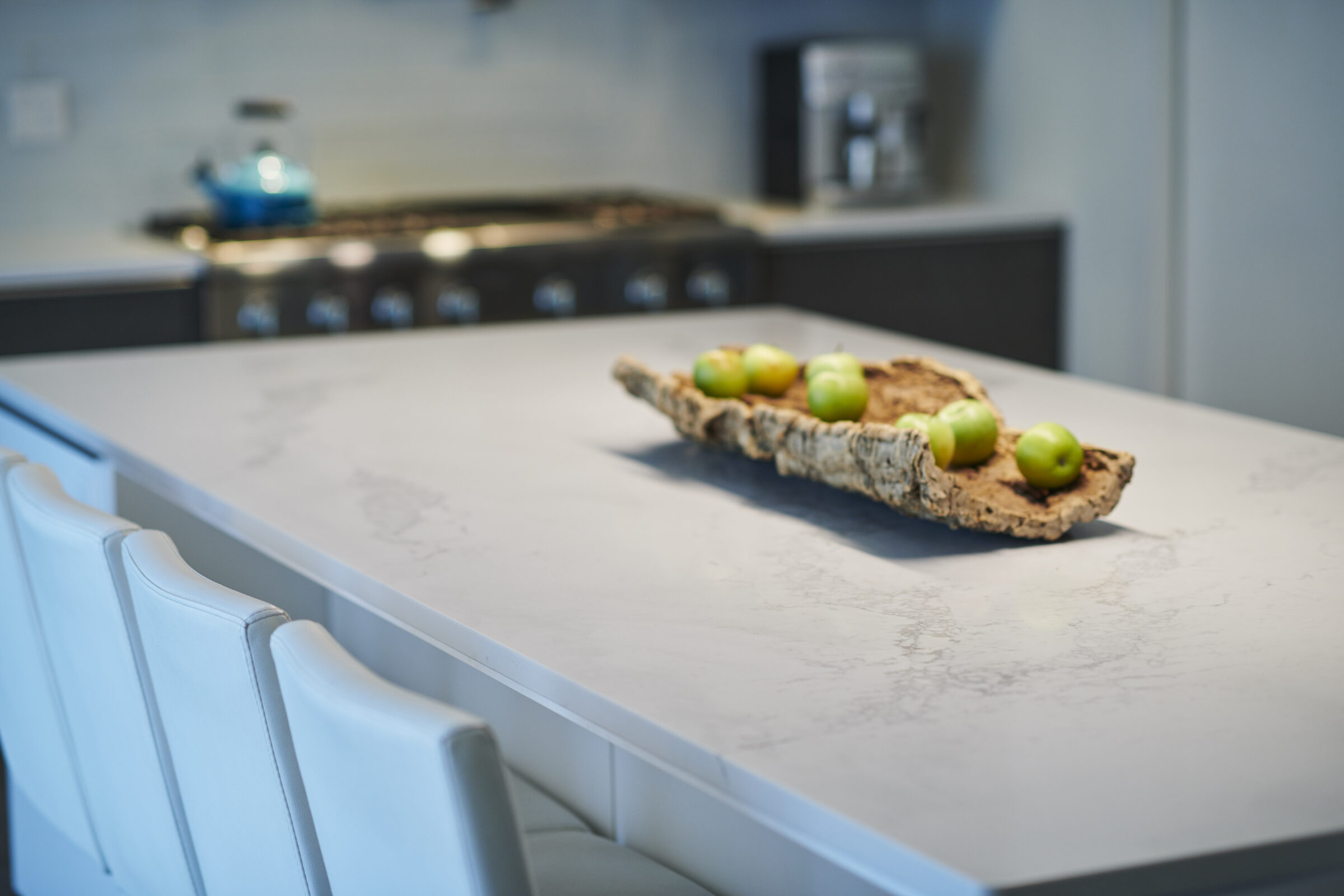 A modern kitchen with a marble island, white chairs, and a wooden centerpiece holding green apples. A stove and cabinets are in the background.