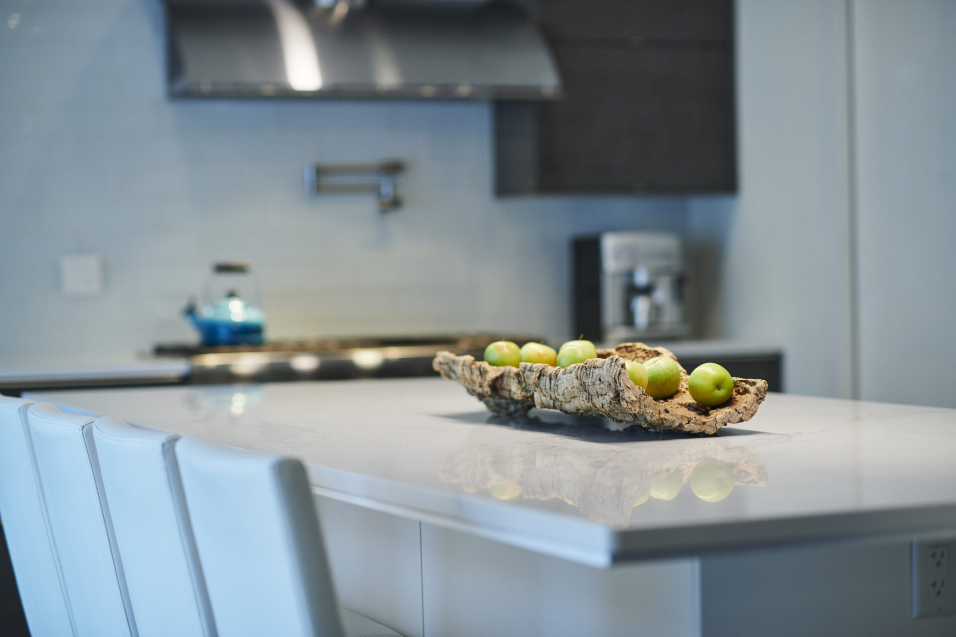 A modern kitchen interior with a white countertop, blurred blue kettle, stainless steel range hood, and a unique wooden fruit tray with green apples.