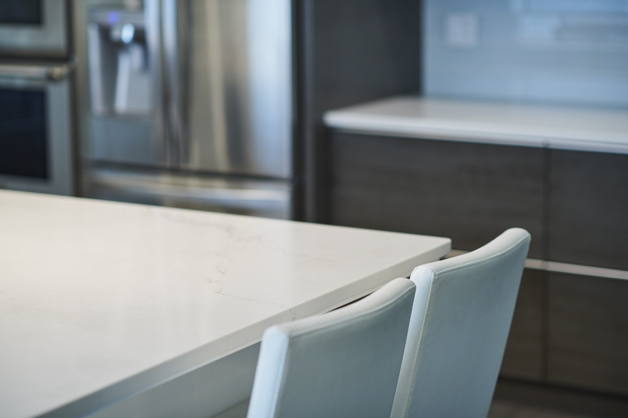 A modern kitchen interior with a white table, light blue chairs, stainless steel refrigerator, and a blurred background emphasizing the eating area.