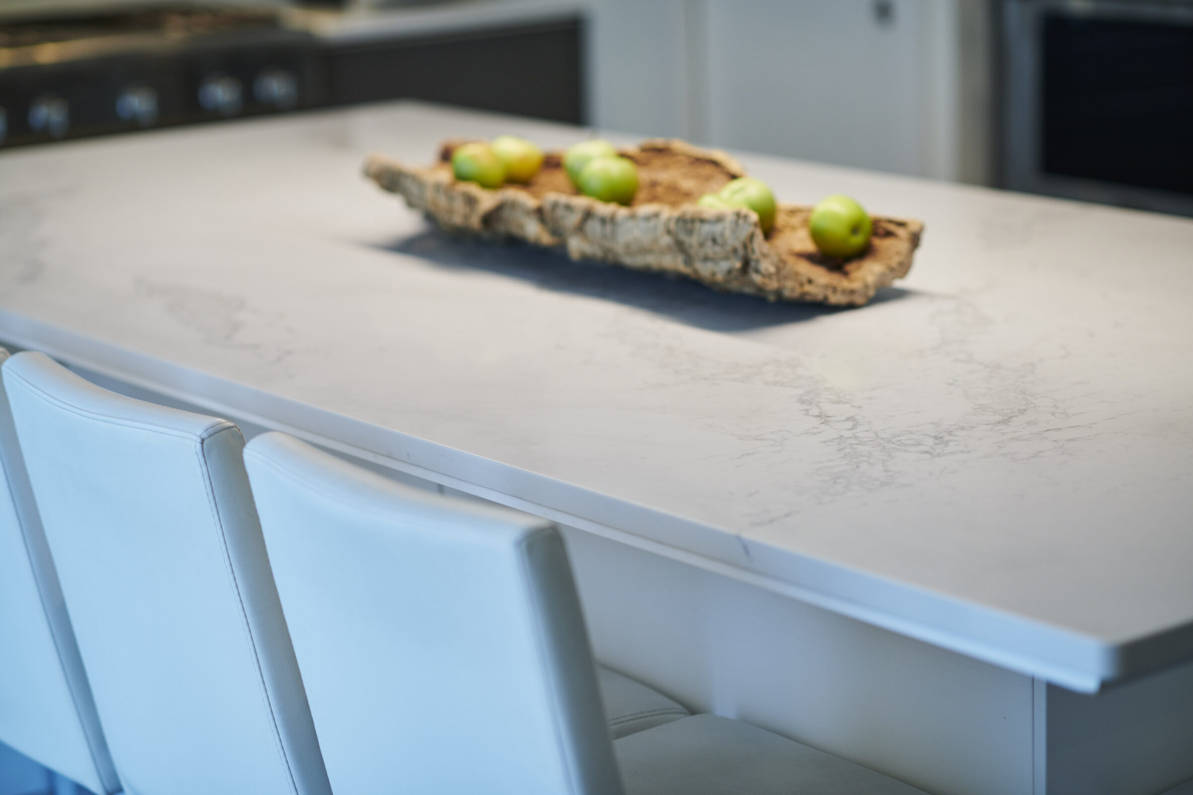 A modern kitchen with a marble countertop, white chairs, and a rustic wooden tray holding green apples. The setting is bright and clean.