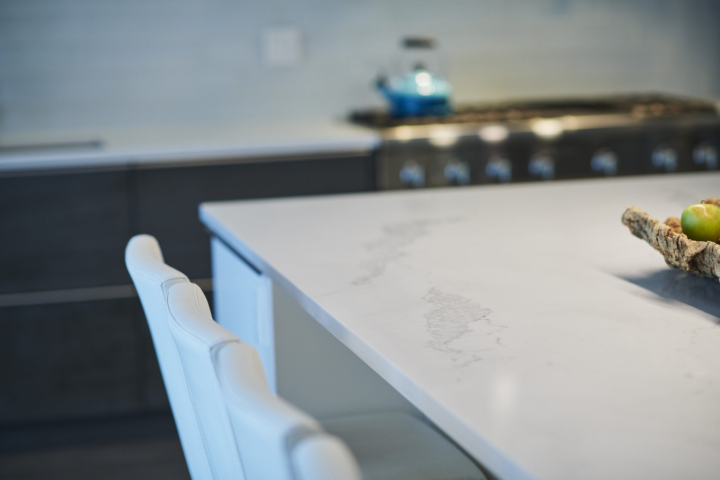 An elegant modern kitchen with a marble countertop, white chairs, stainless steel stove, blue kettle, and an apple on a textured mat.