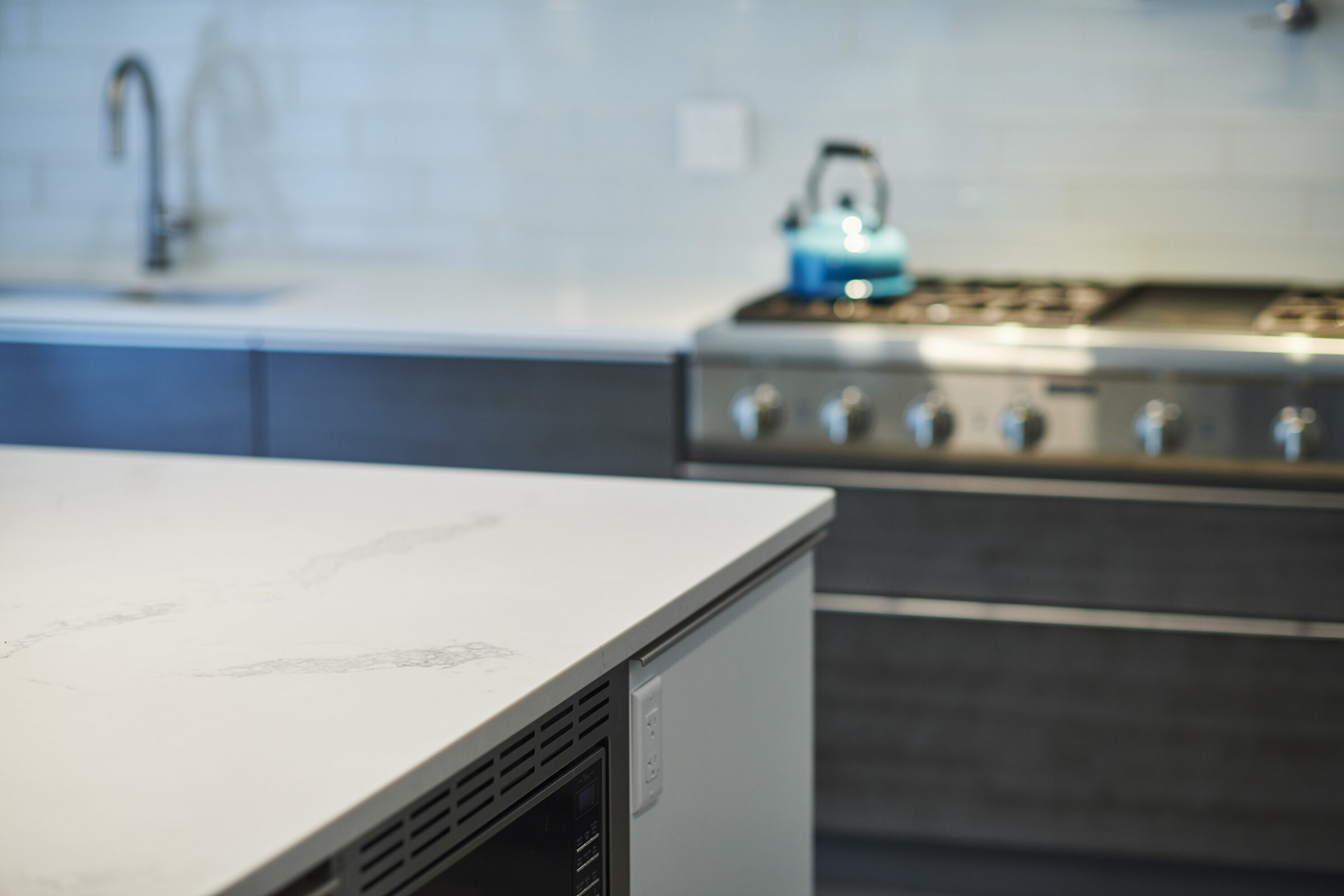 A modern kitchen with stainless steel appliances, white countertops, and a blue kettle. Focus is on the foreground with a blurred background.