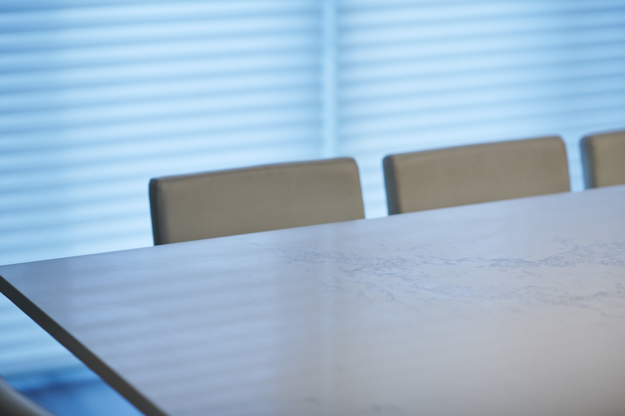 This image shows a modern meeting room with a sleek marble table and three empty chairs, against a backdrop of closed horizontal window blinds.