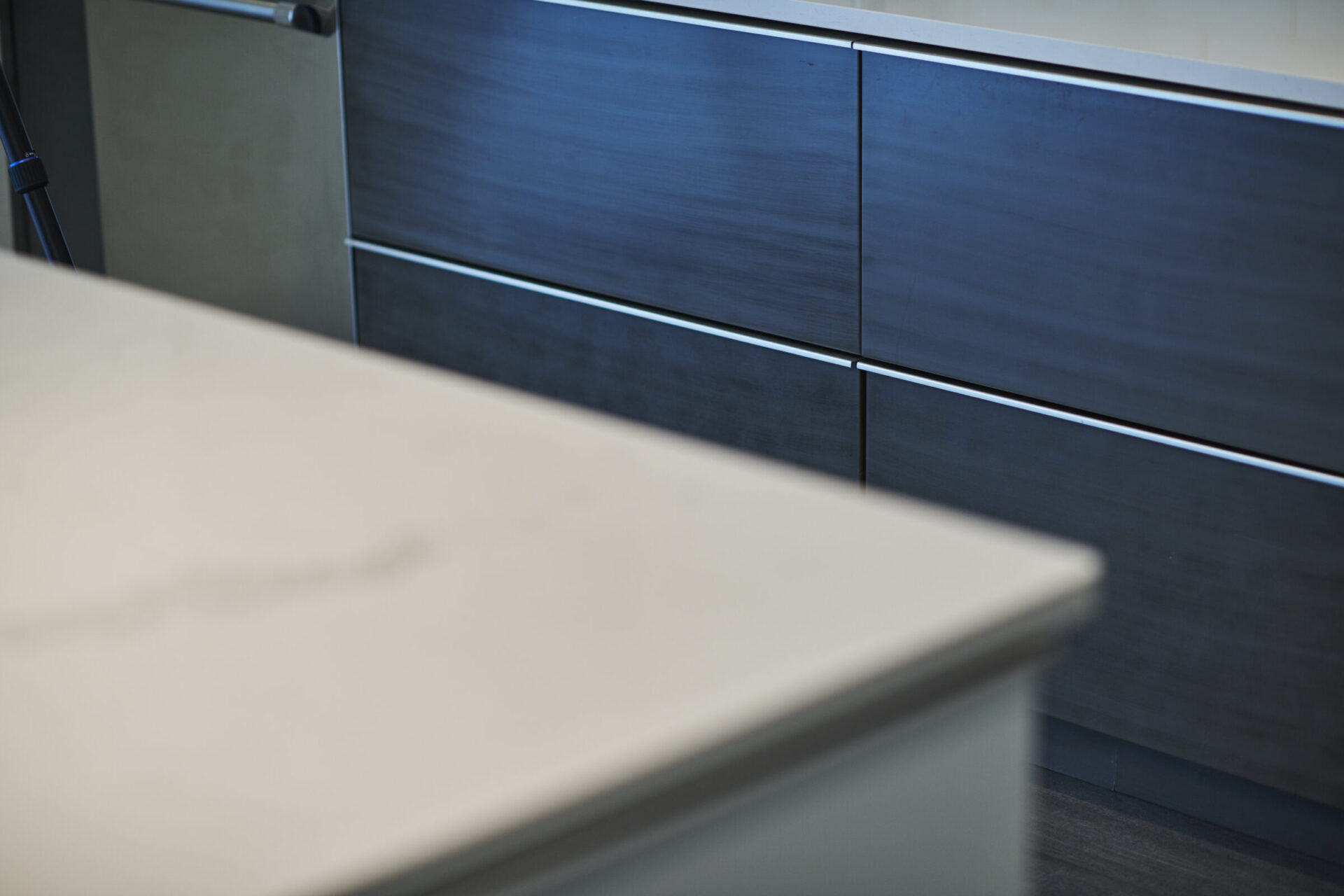 Close-up, angled view of a modern kitchen showing a corner of a beige countertop with sleek, dark cabinetry and silver handles in the background.