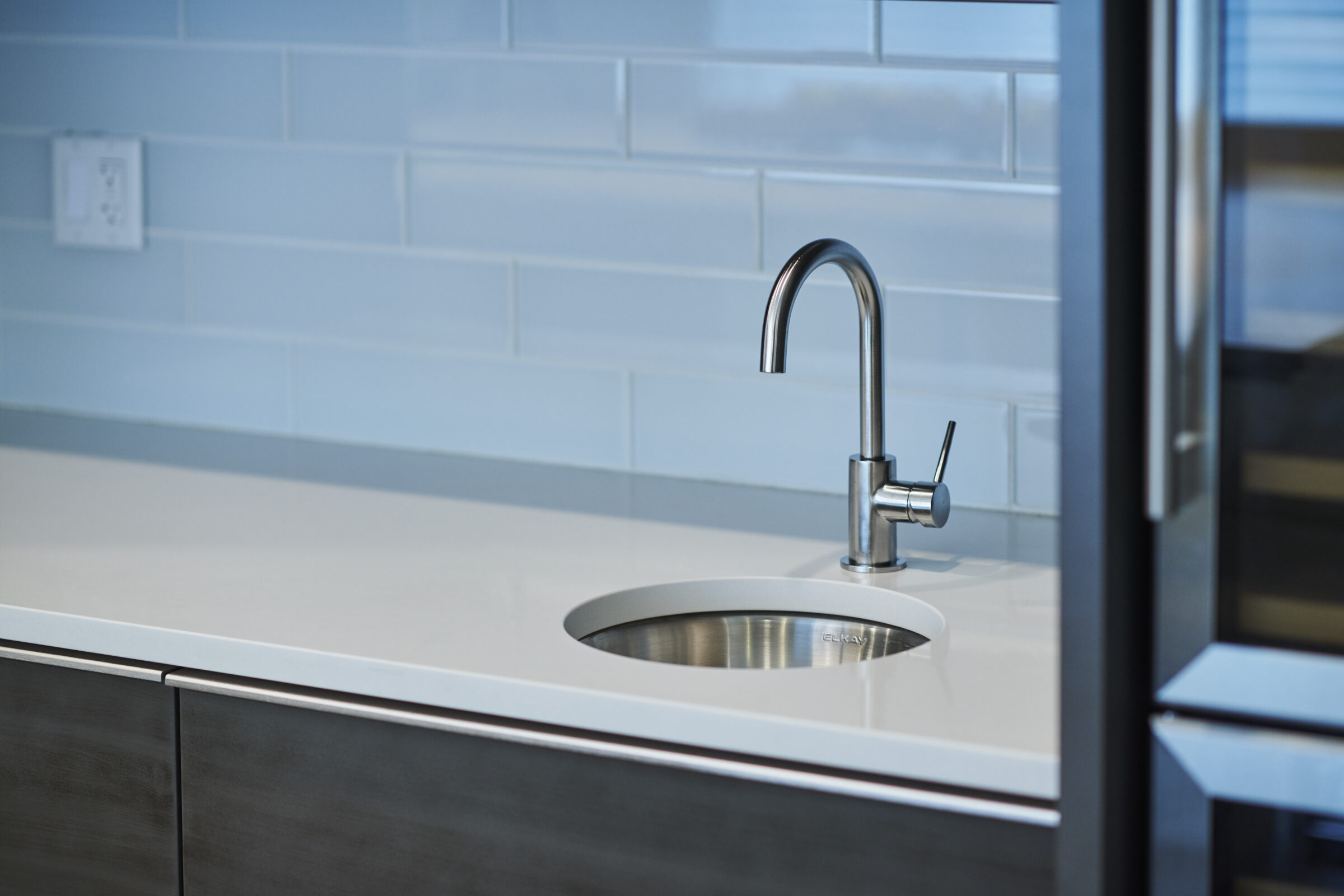 A modern kitchen with a stainless steel sink and faucet against a blue tile backsplash. White countertop, dark cabinets, and no people visible.