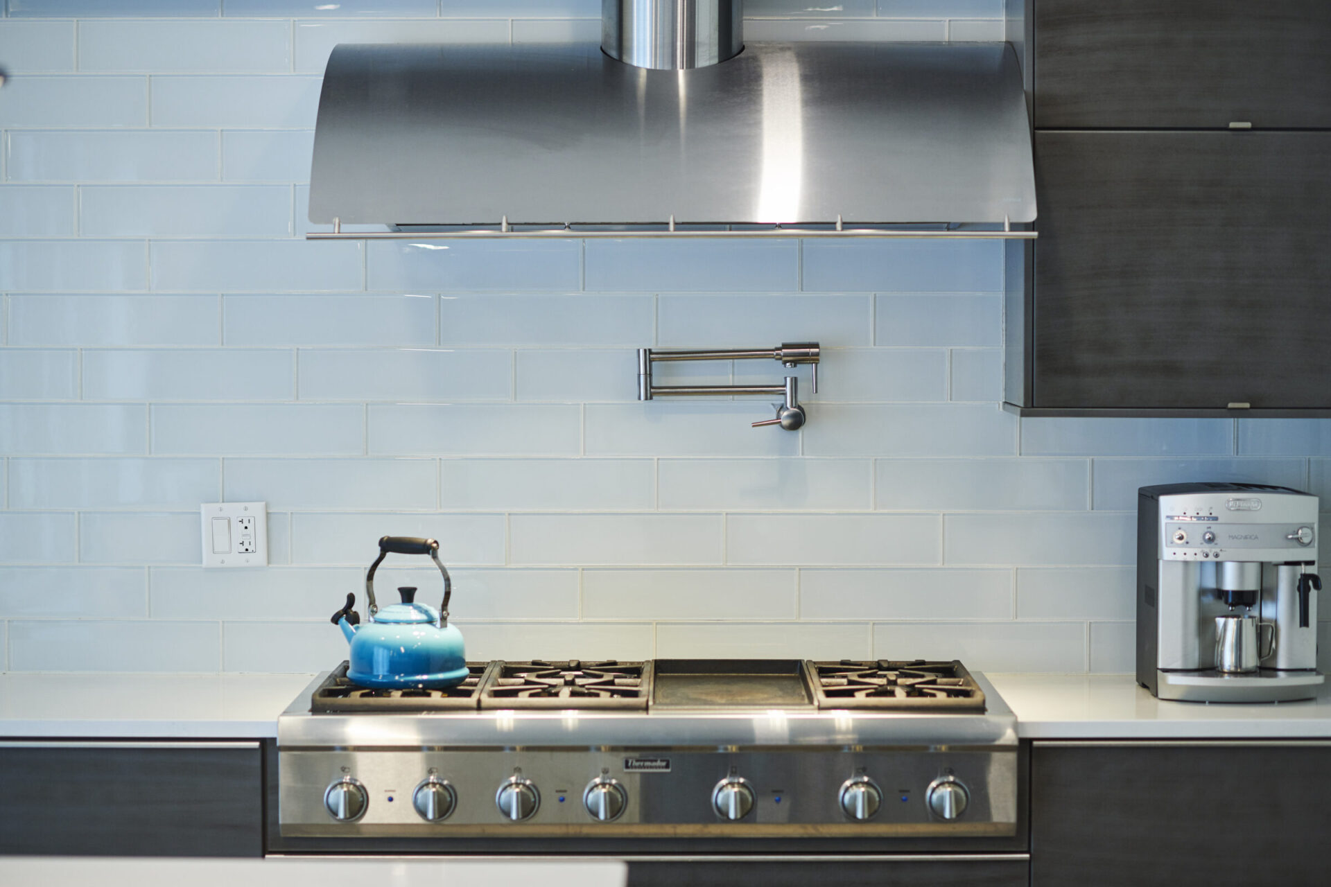 A modern kitchen with stainless steel appliances, a blue kettle on the stove, light blue backsplash tiles, and a sleek coffee machine.