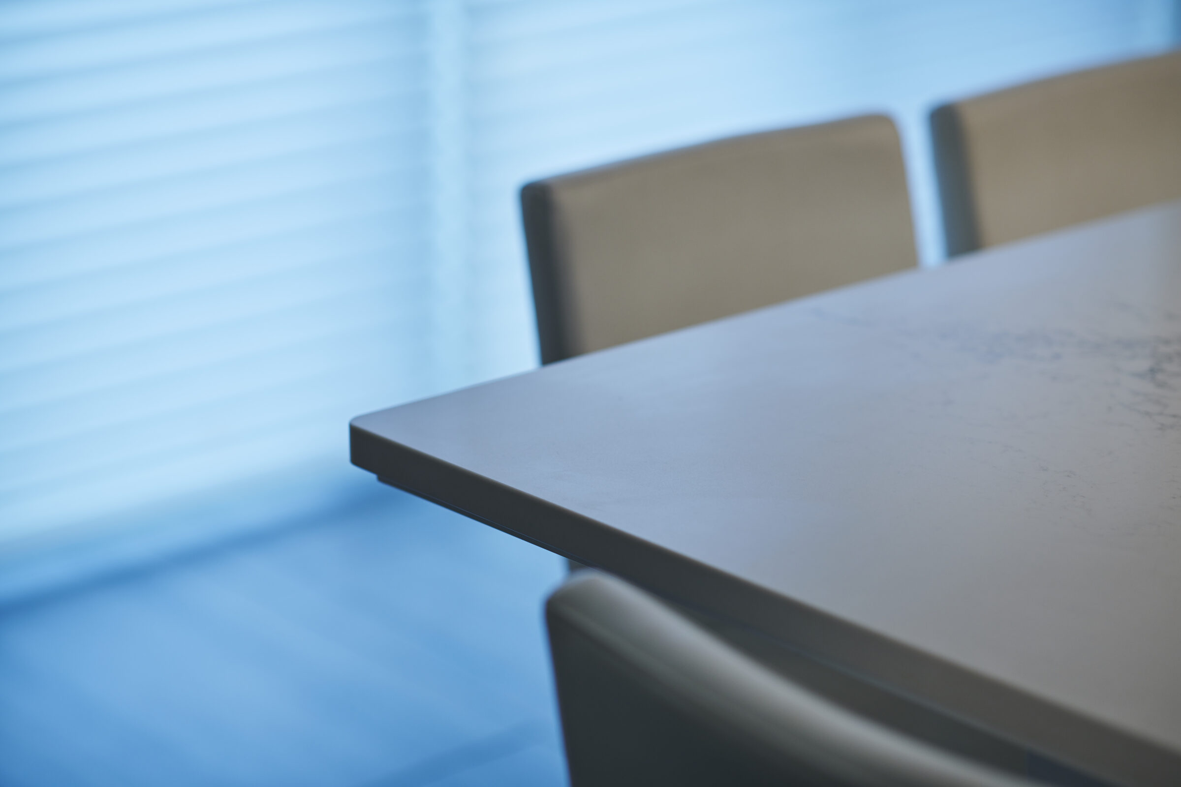 A corner of a modern table with a smooth surface is shown with chairs against a blue backdrop, possibly in an office meeting room.