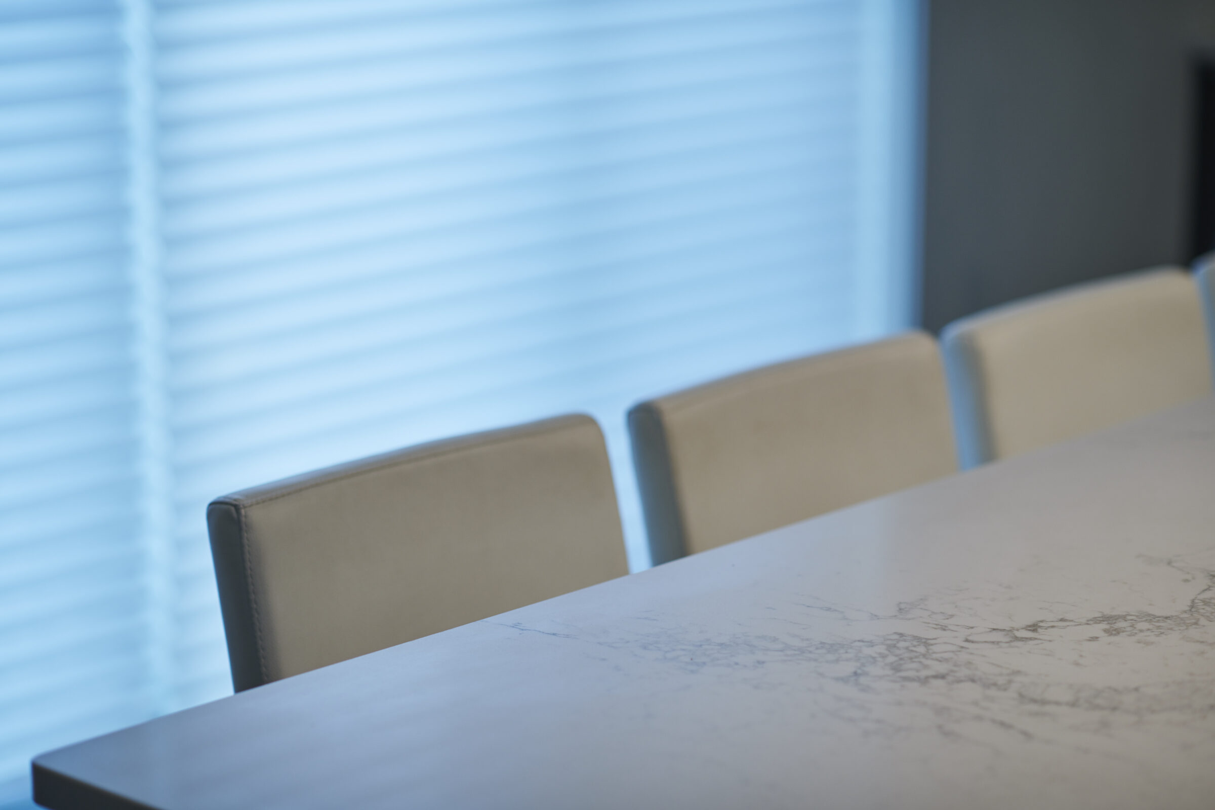This image shows a modern meeting room with a marble table and four beige chairs against a backdrop of closed horizontal window blinds.