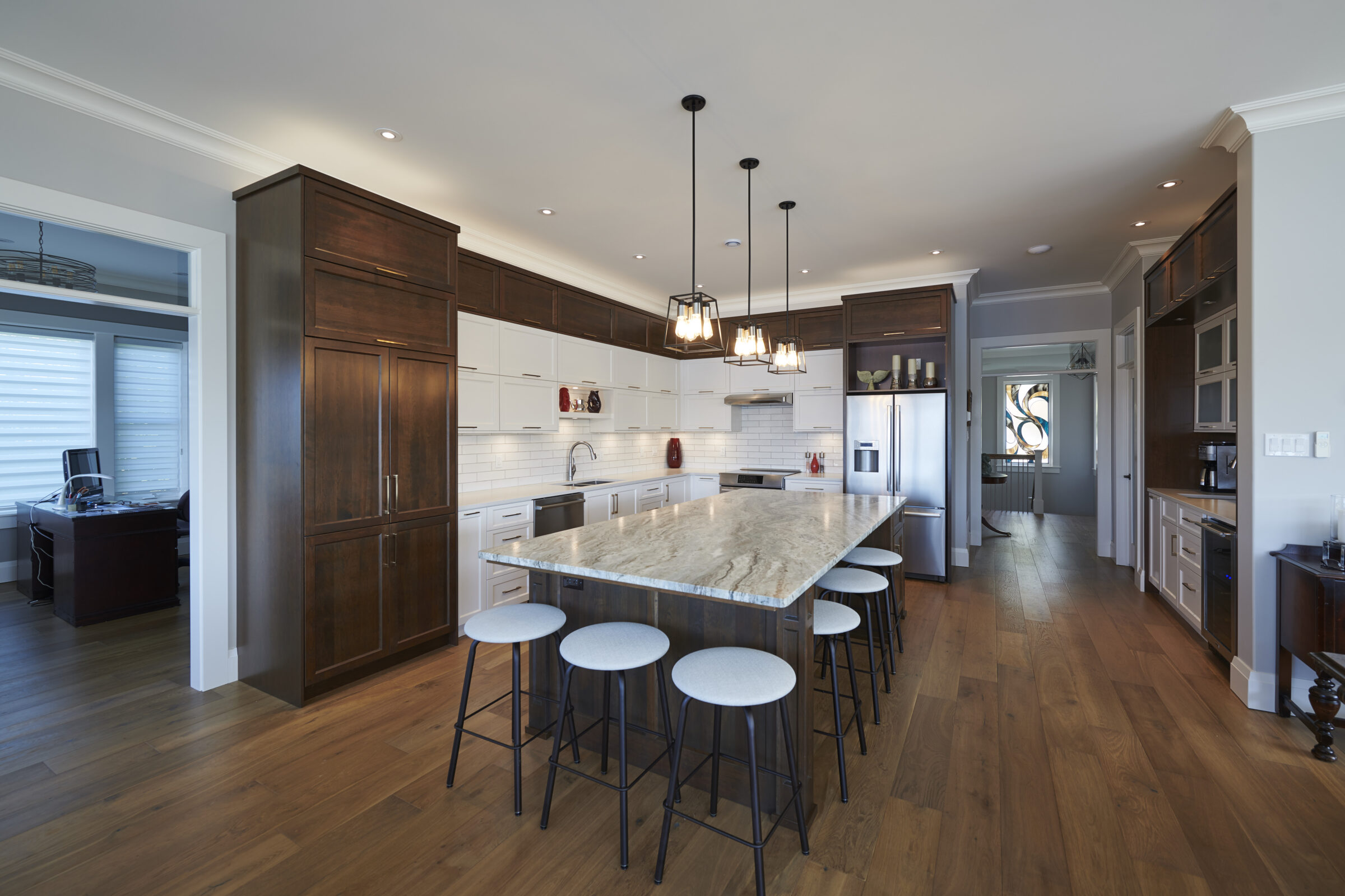 A spacious, modern kitchen with dark wood cabinets, white backsplash, a large island with bar stools, stainless steel appliances, and pendant lights.