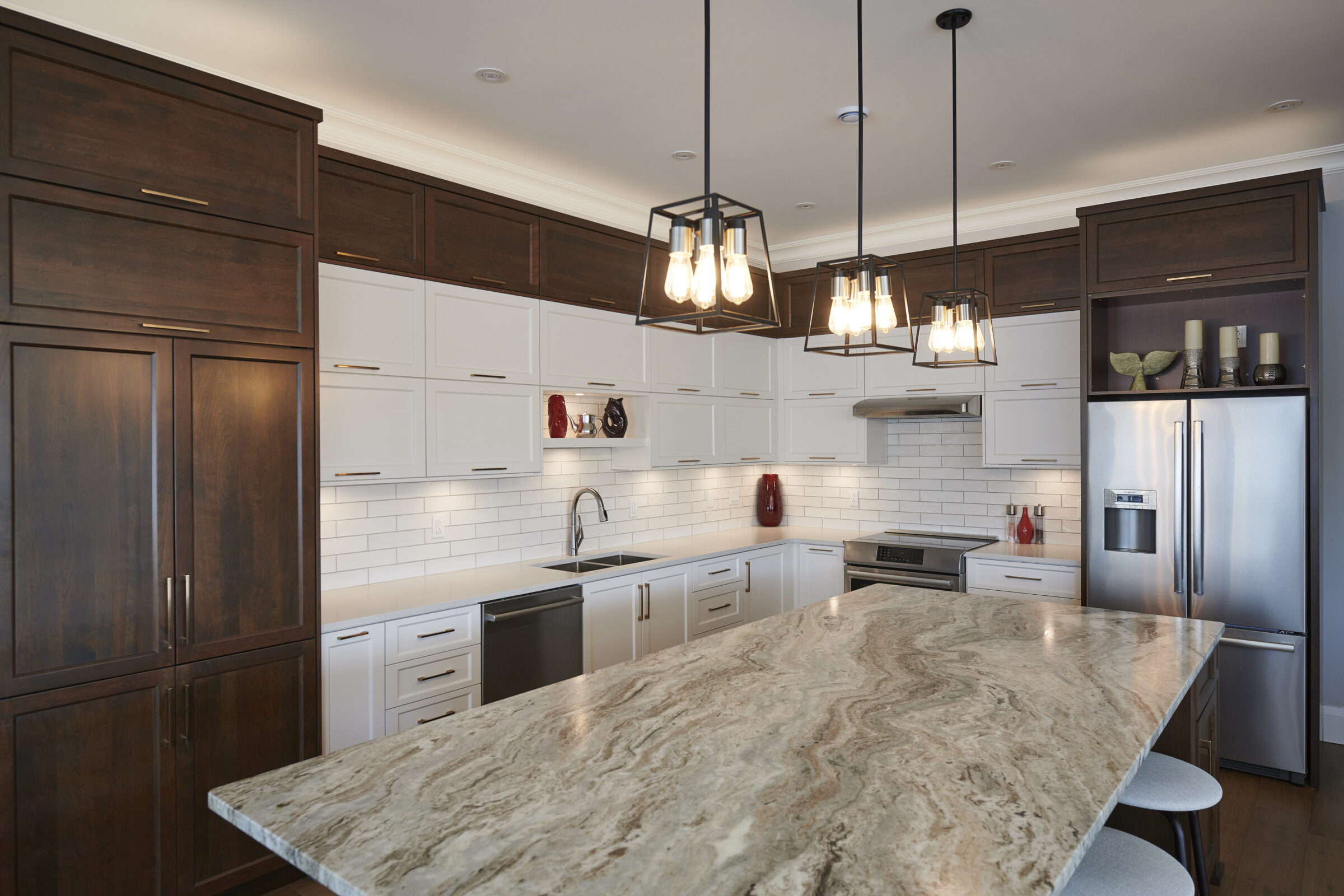A modern kitchen with white cabinetry, dark wood accents, stainless steel appliances, pendant lights, and a large island with a marble countertop.