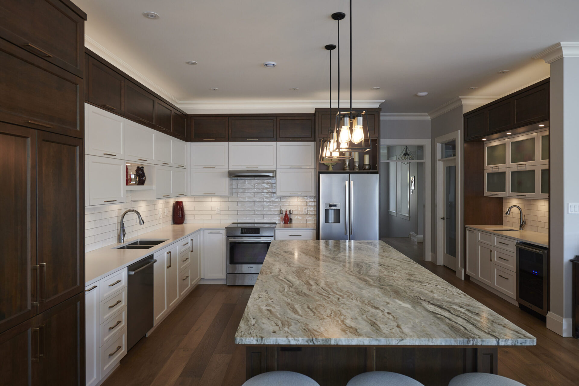 Modern kitchen with white cabinets, dark wood trim, stainless steel appliances, subway tile backsplash, hanging lights, and a large marble-topped island.
