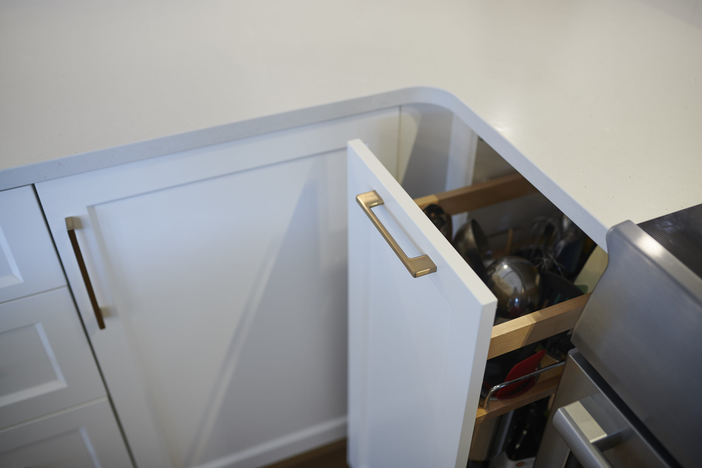 A corner kitchen cabinet with a white door partially open, revealing wood shelves storing assorted pots and pans, against a stone countertop.