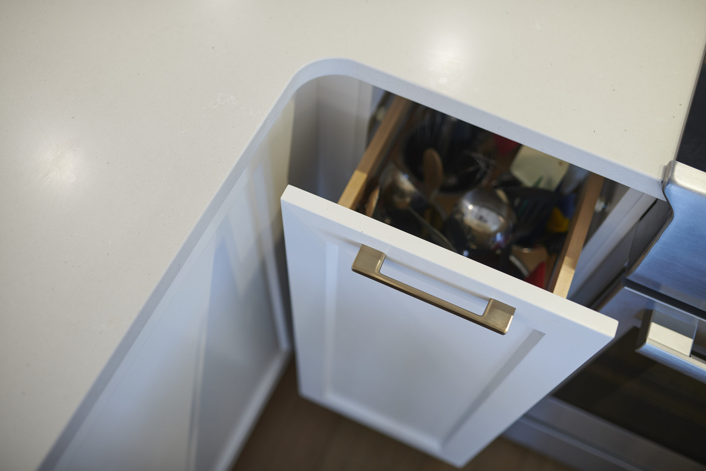 An open kitchen drawer is filled with various utensils. It has a white front with a silver handle, under a beige countertop.
