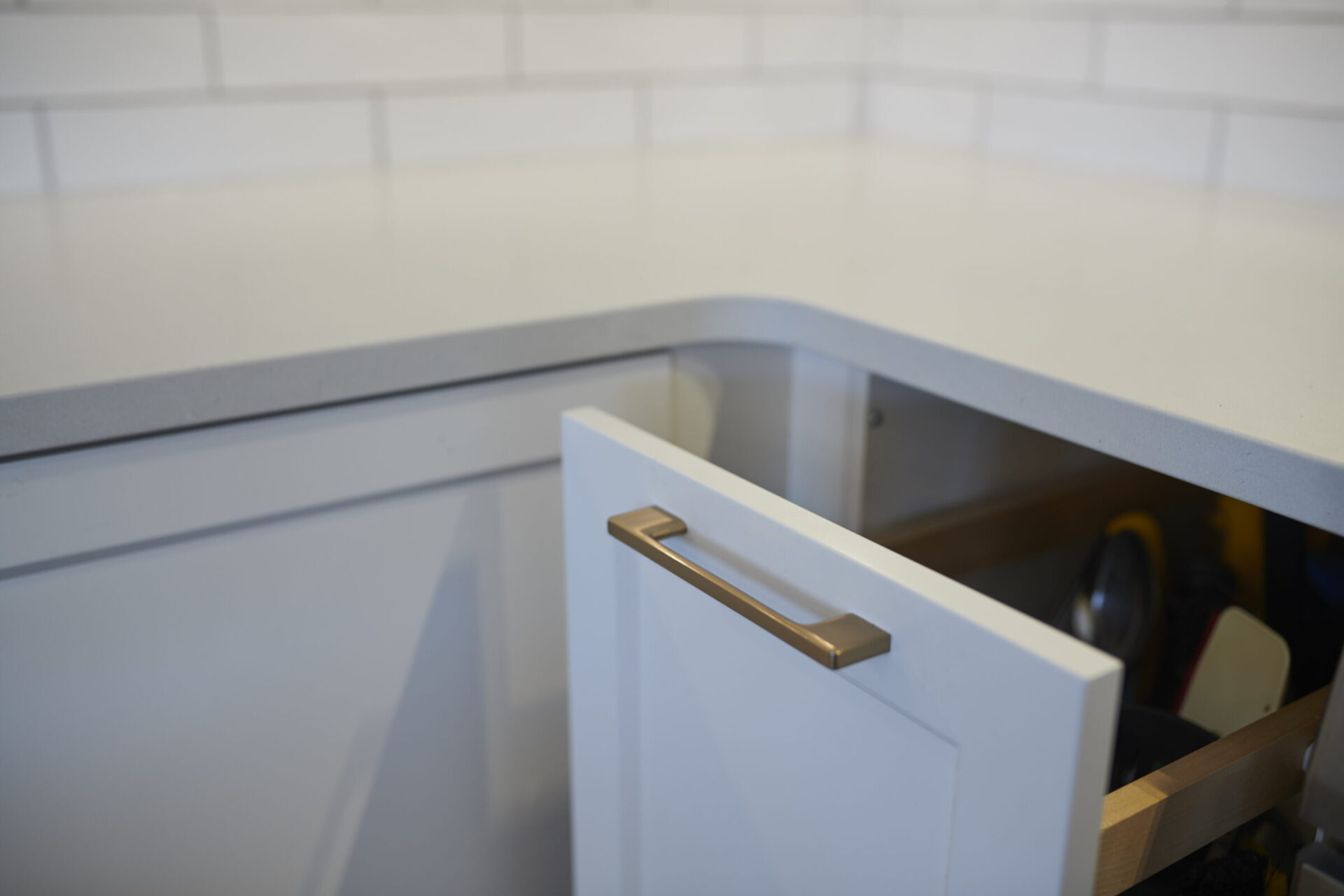 This image shows a partially opened white drawer in a modern kitchen, revealing a glimpse of pots and pans inside and a tiled backsplash above.