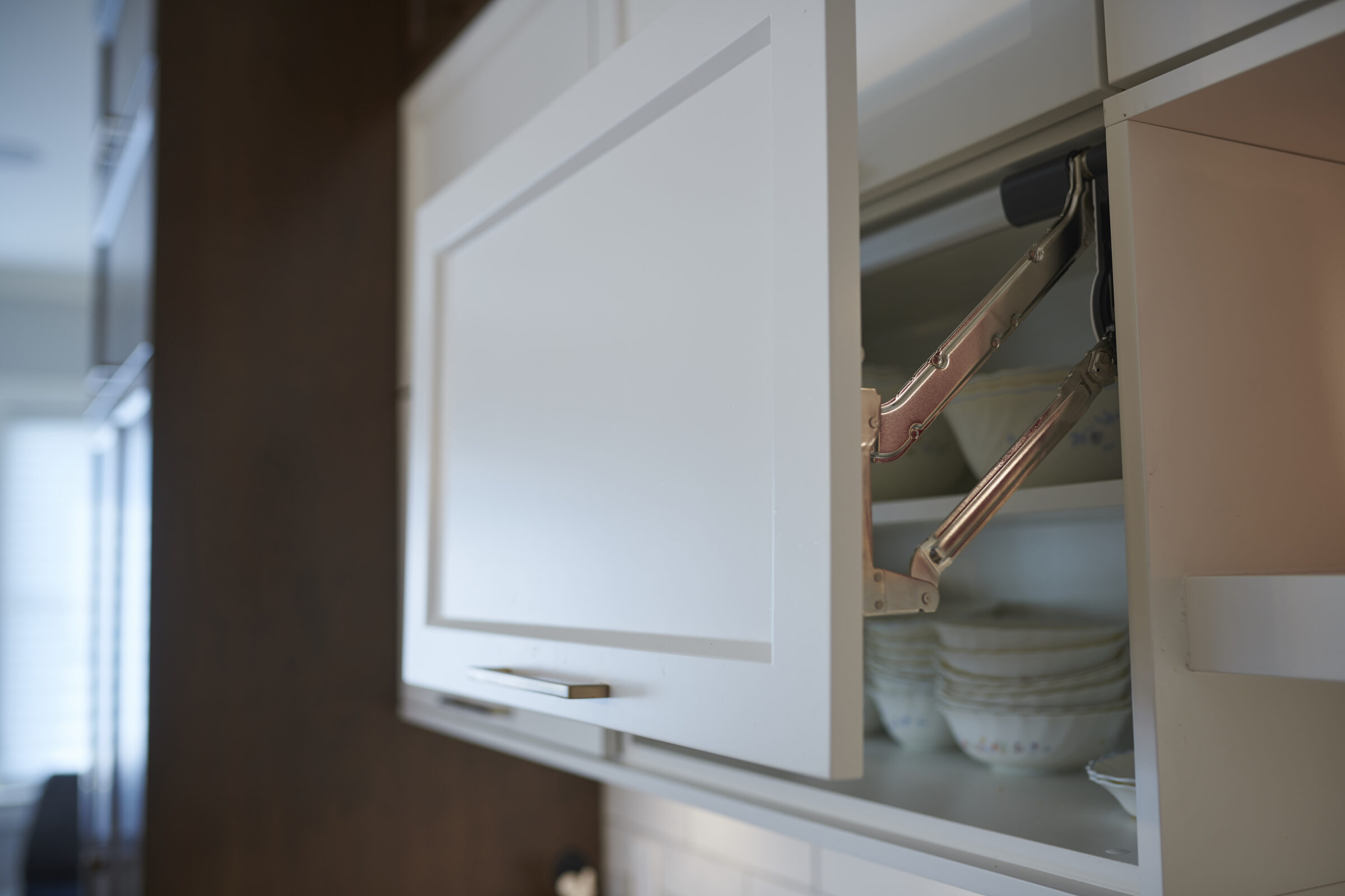 An open cabinet with a hydraulic arm, showcasing stacked plates inside. The kitchen environment appears modern and clean with soft lighting.
