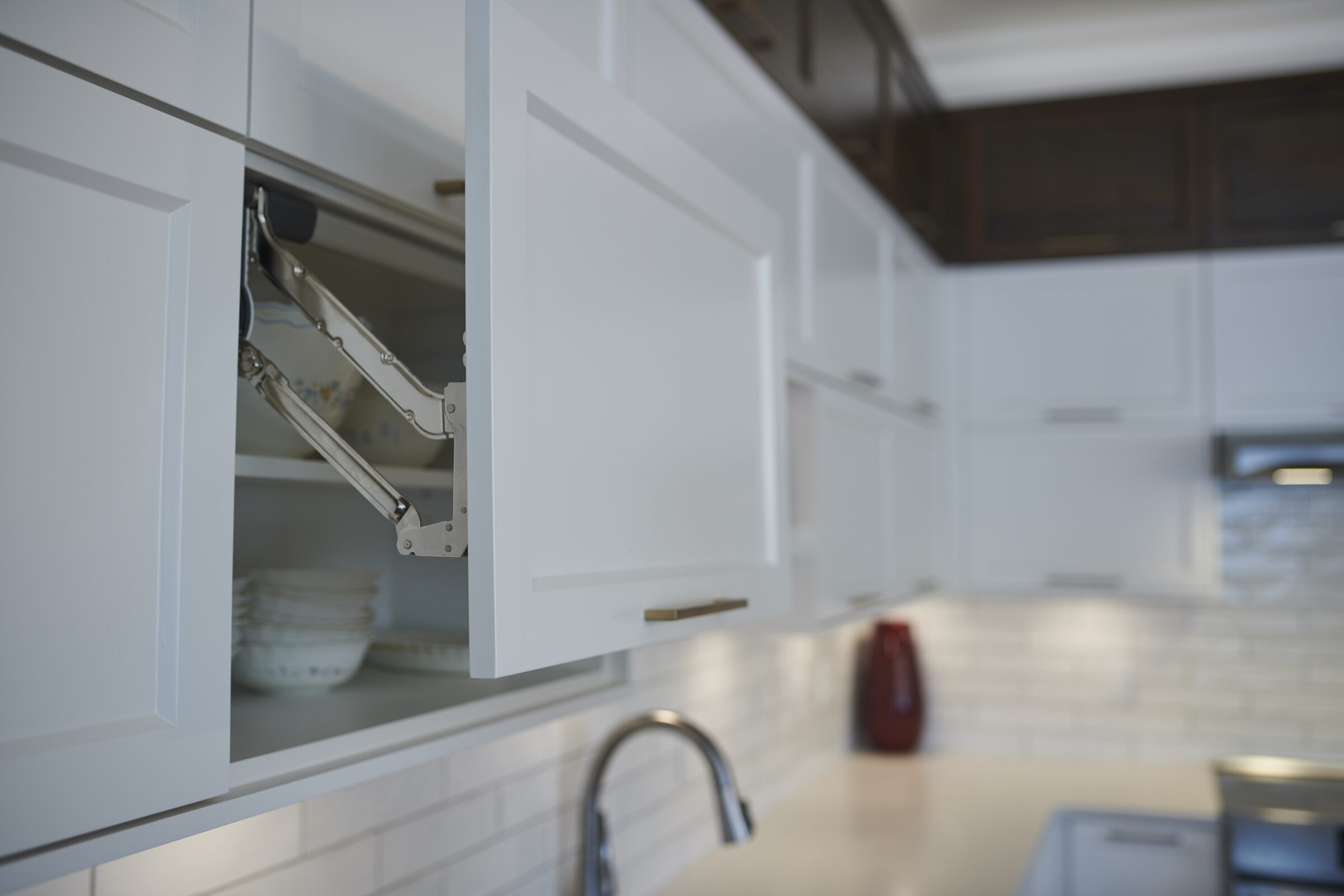 A modern kitchen interior with white cabinetry, a lifted cabinet door with a mechanical hinge, subway tile backsplash, and a stainless steel sink.