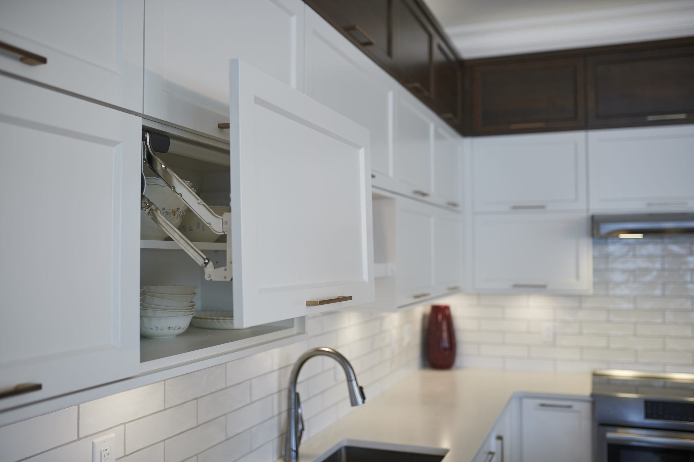 A modern kitchen with white cabinetry, a lifted cabinet door revealing stacked dishes, stainless steel appliances, and a subway tile backsplash.
