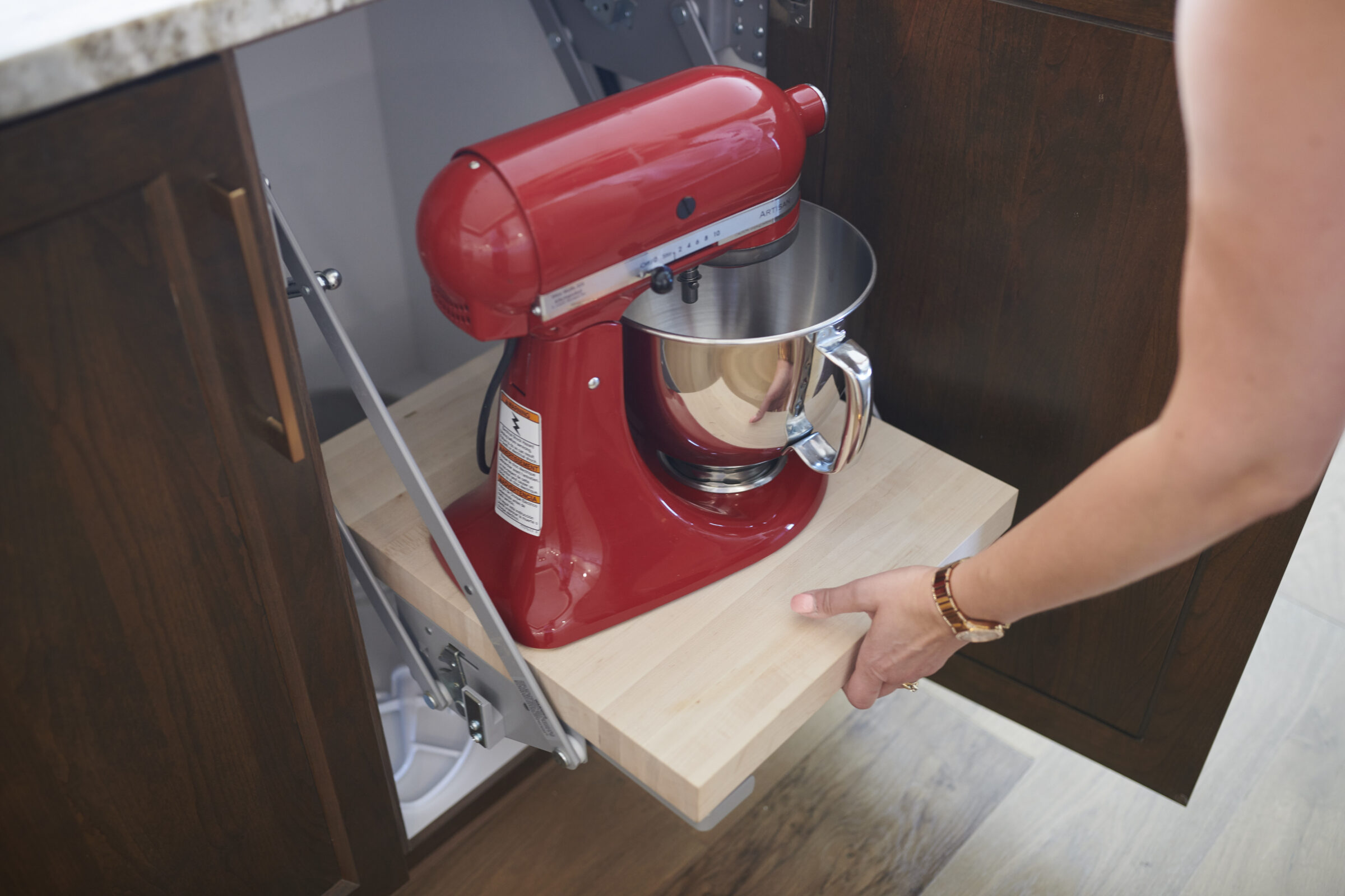 A person is pulling out a shelf with a red stand mixer from a dark wood kitchen cabinet, showcasing a smart storage solution.