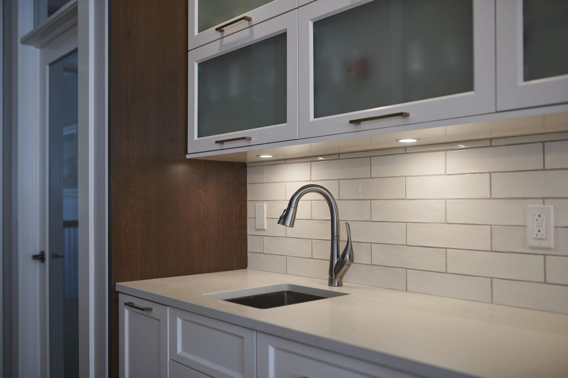 A modern kitchen with white subway tile backsplash, stainless steel faucet, and dark wood cabinets alongside white countertops and LED under-cabinet lighting.