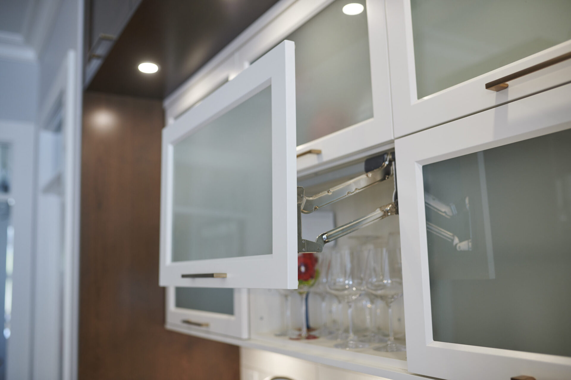 An open white kitchen cabinet with frosted glass doors, revealing stemware glasses inside. The cabinet features modern hardware and a softly lit interior.