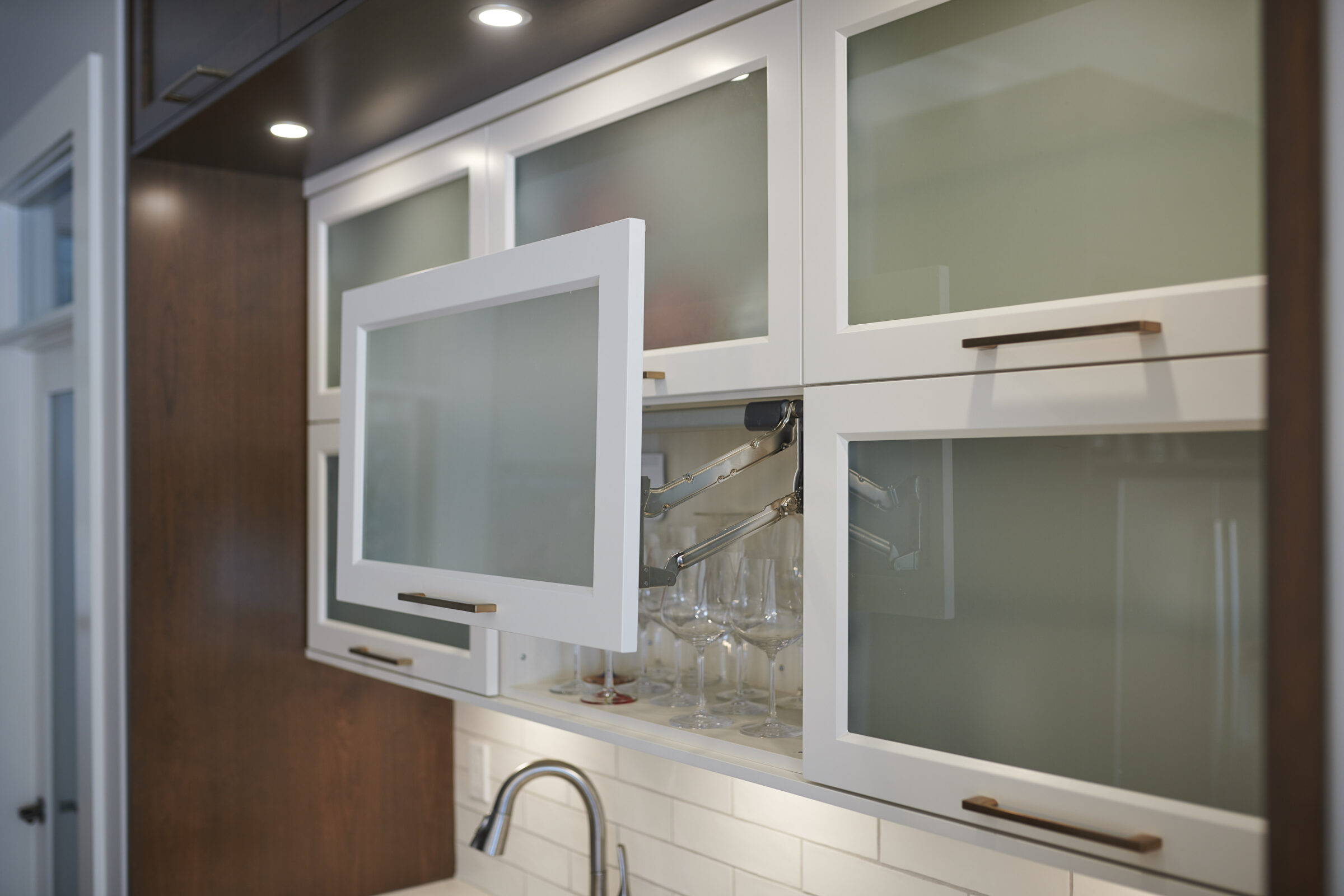 Modern kitchen cabinet with frosted glass doors open, revealing stemware inside. White tile backsplash and dark wood visible, with a sleek design aesthetic.