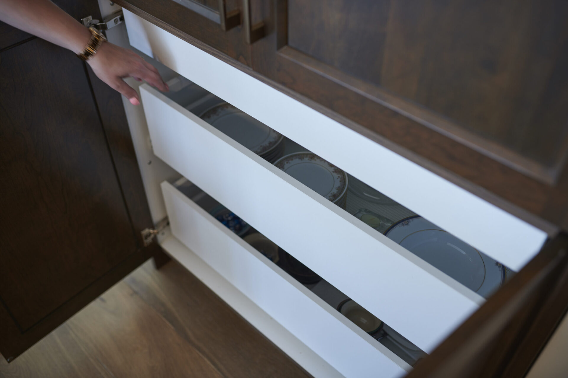 A person is opening a modern, dark wood finish cabinet drawer containing neatly arranged plates. The focus is on the drawer's contents and the person's hand.